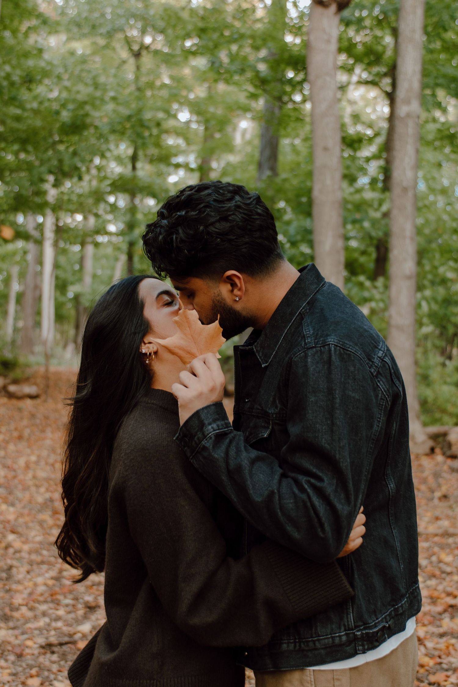 A couple shares a romantic almost-kiss moment in an autumn forest, surrounded by tall trees and fallen leaves.