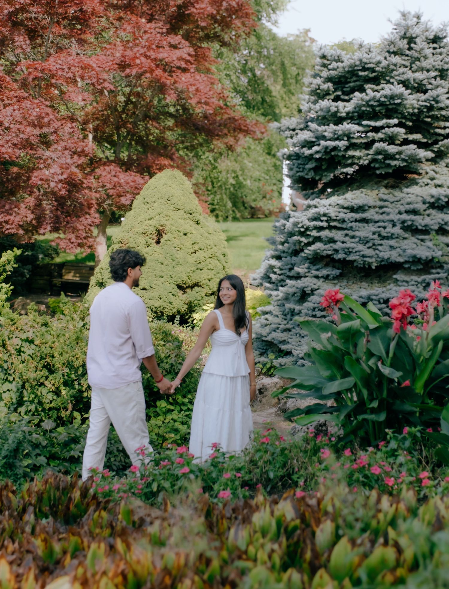 A couple holds hands while walking through a lush garden with colorful trees and flowers.