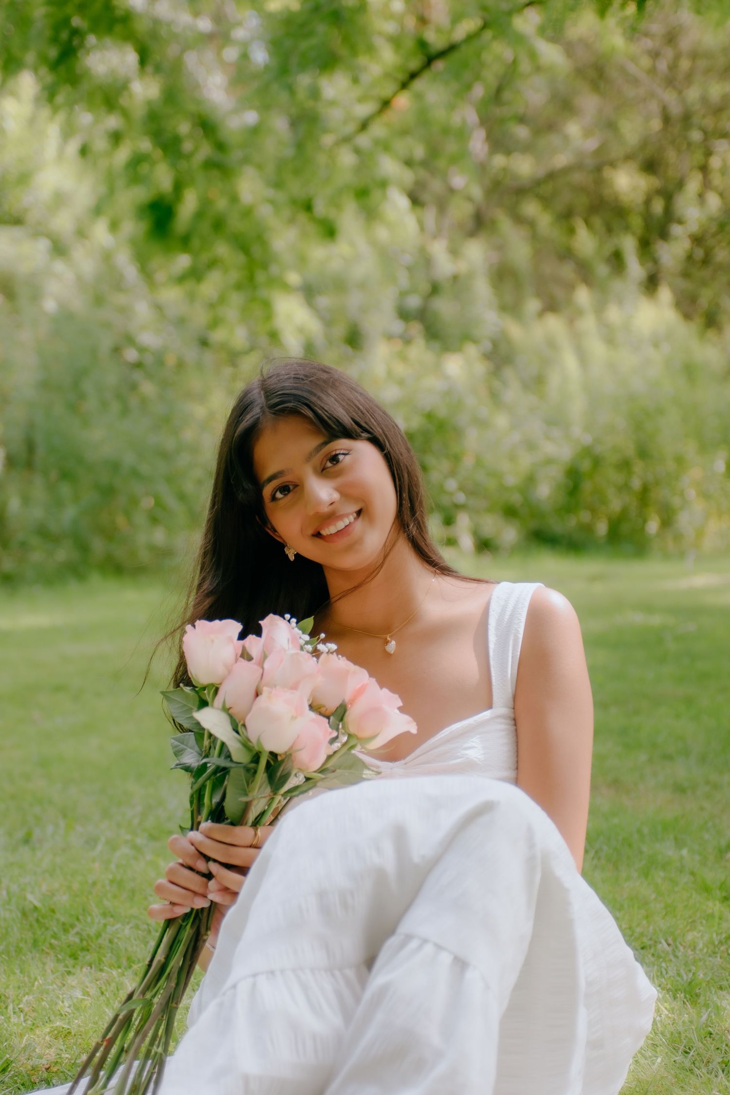 Young woman in white dress sitting on grass holding pink roses bouquet, smiling outdoors in green park setting.