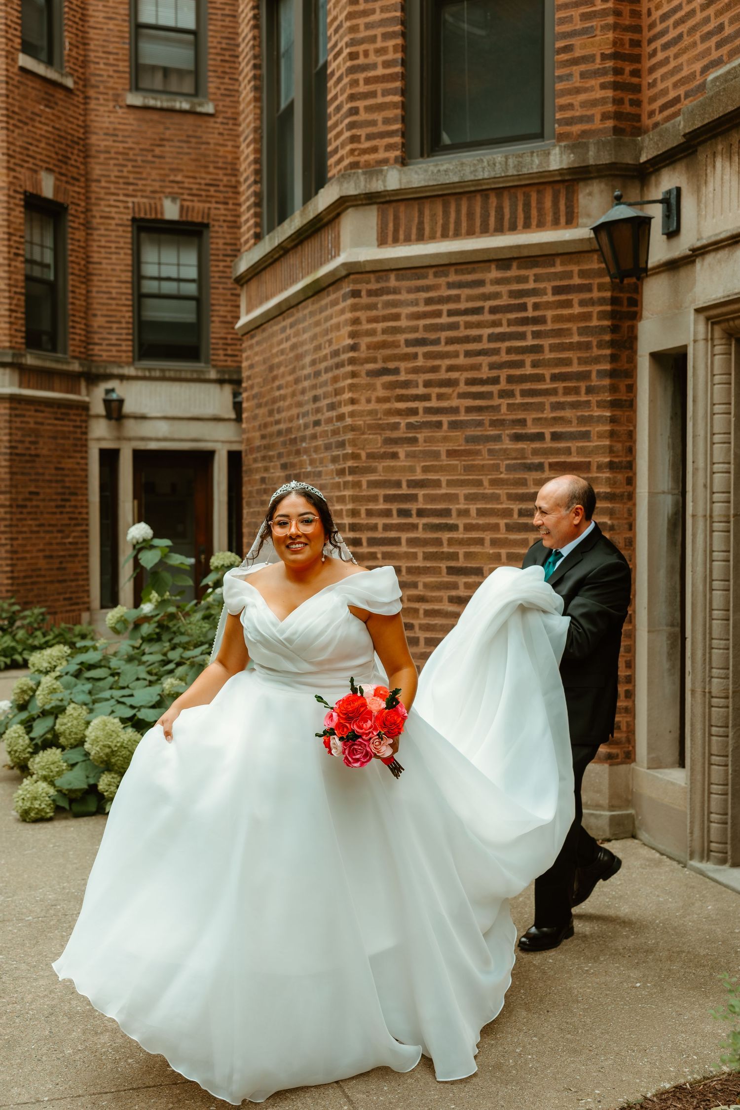 Smiling bride in white ball gown holds red bouquet outdoors as man carries her dress train behind her. Chicagoland weddings.