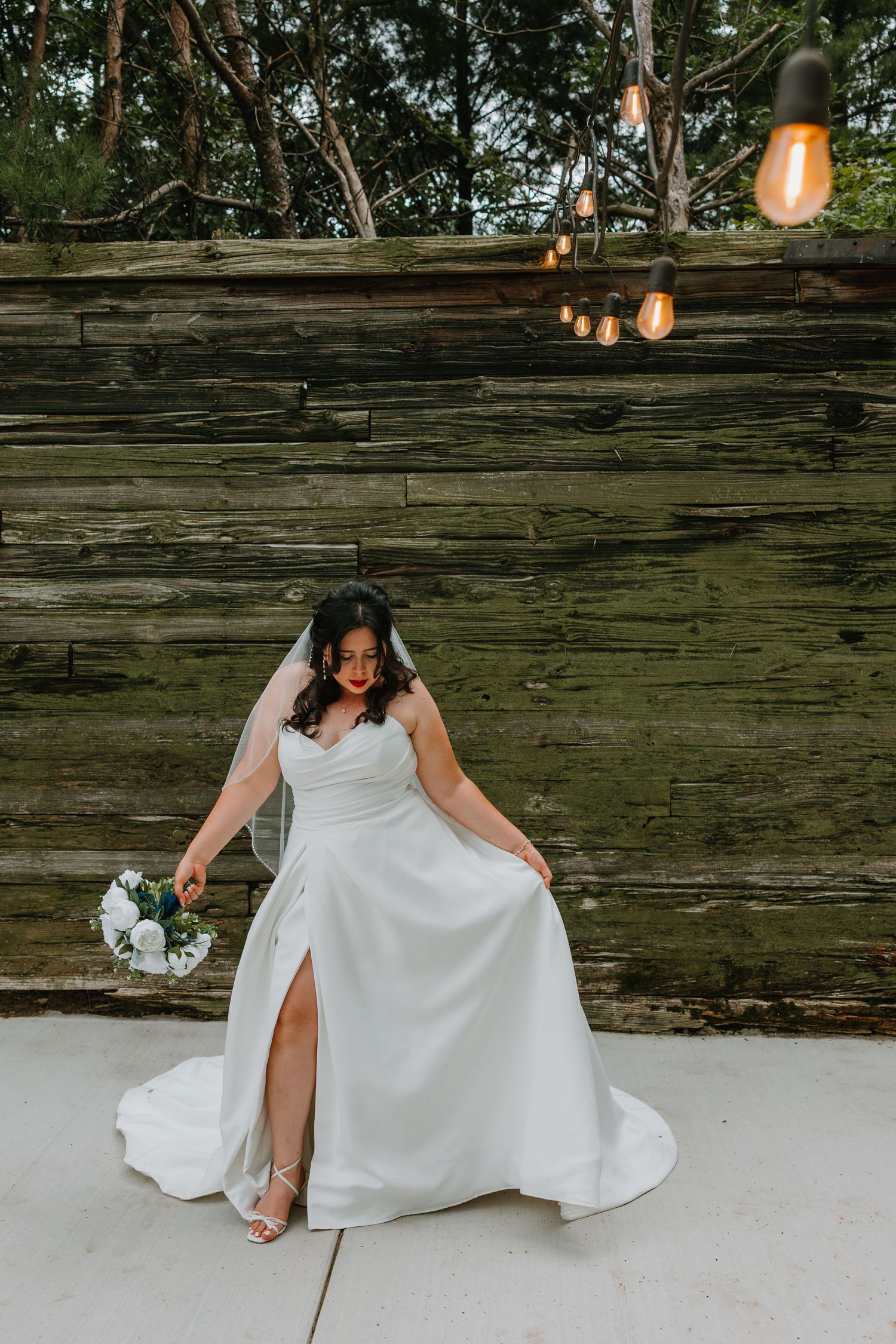 Elegant bride in a white satin gown with a thigh slit poses with bouquet against a rustic wooden wall. Northwest Indiana wedding venues.