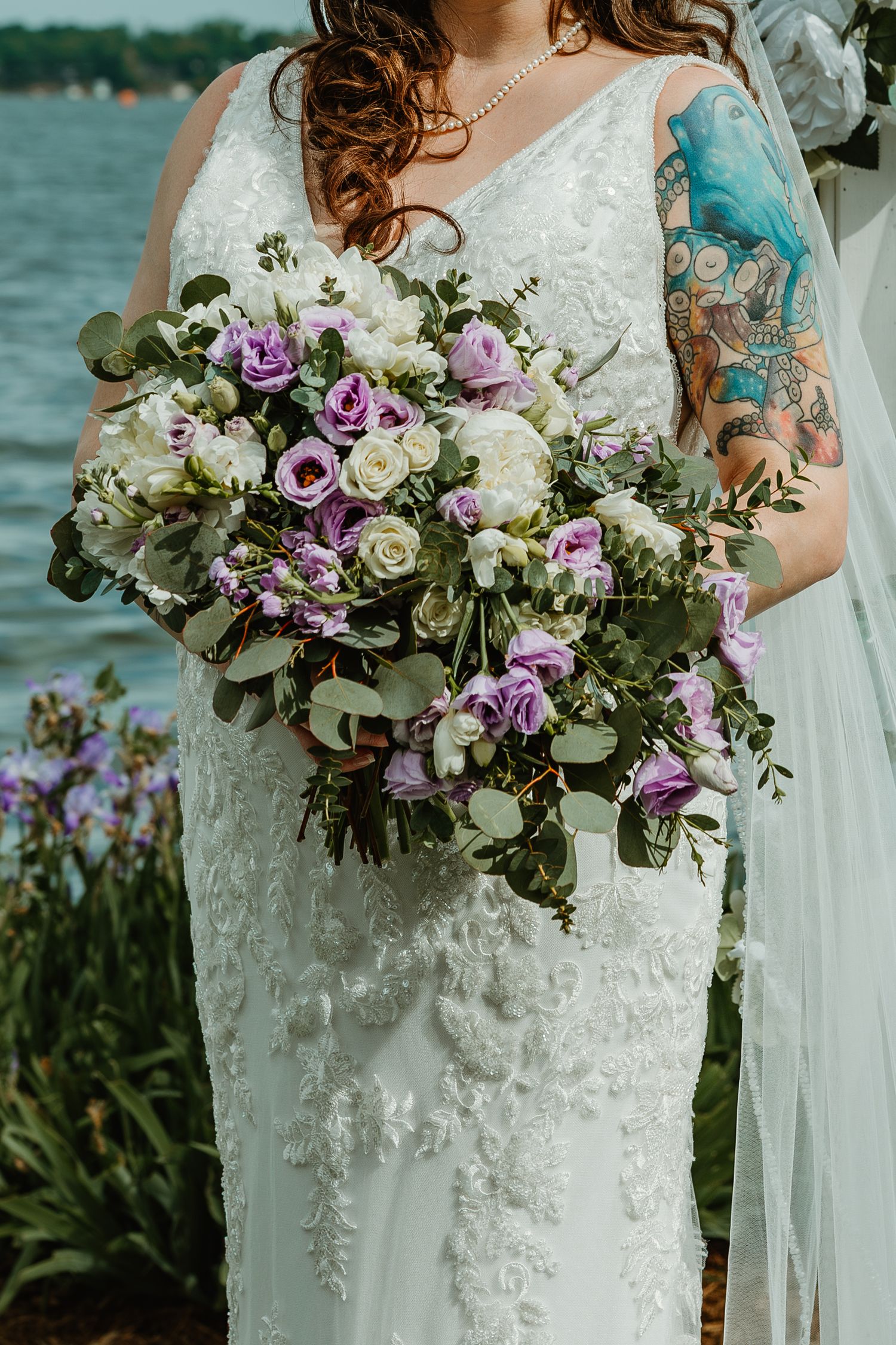 Close-up of tattooed bride holding lush purple and white rose bouquet in lace wedding dress by lake.