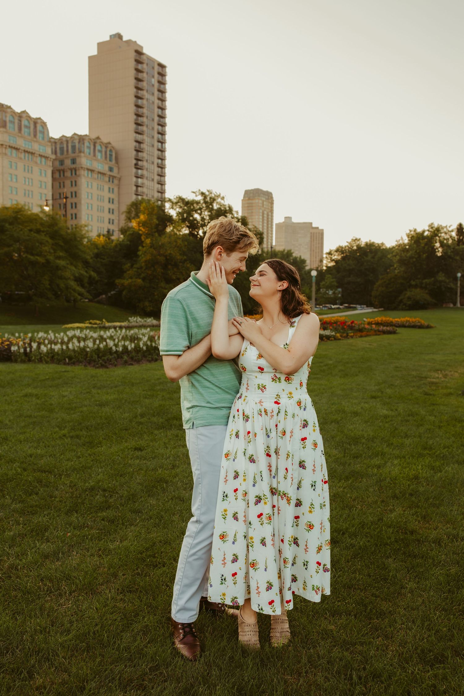 Couple sharing a tender moment on a green lawn, woman cradling man's face with city skyline behind them. Chicago engagement photos.