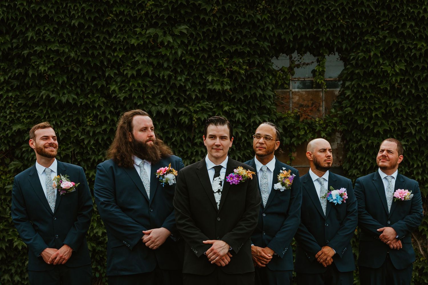Six groomsmen in navy suits with colorful boutonnieres posing in front of ivy-covered wall. Chicagoland wedding venues.