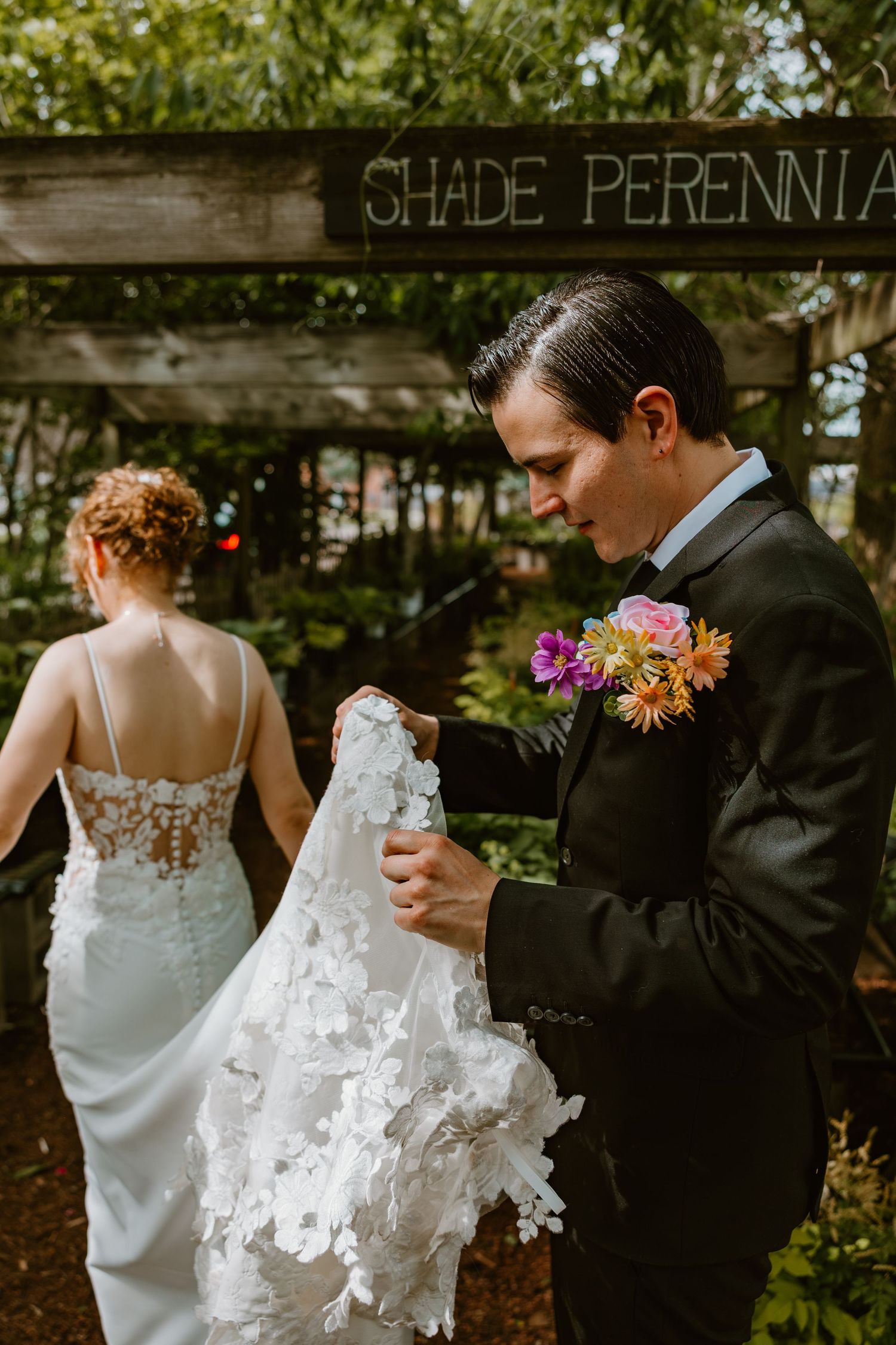 Groom in black suit carries bride's floral lace veil train near shade perennials sign. Chicagoland wedding venues.