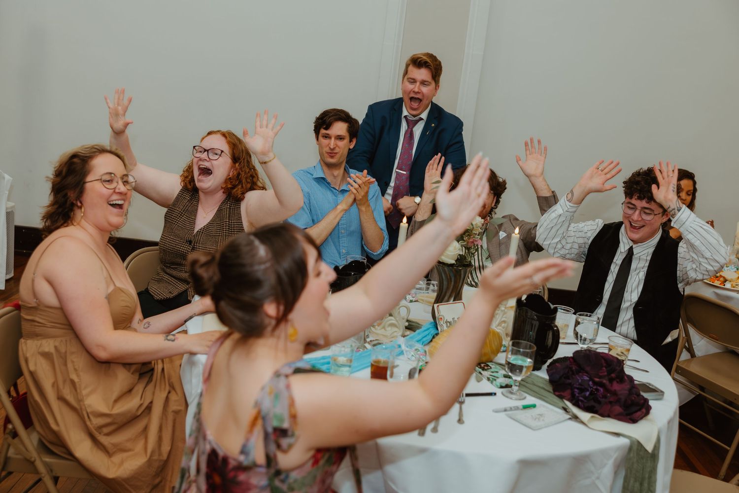 Joyful wedding reception guests raising hands and celebrating together around a decorated table.