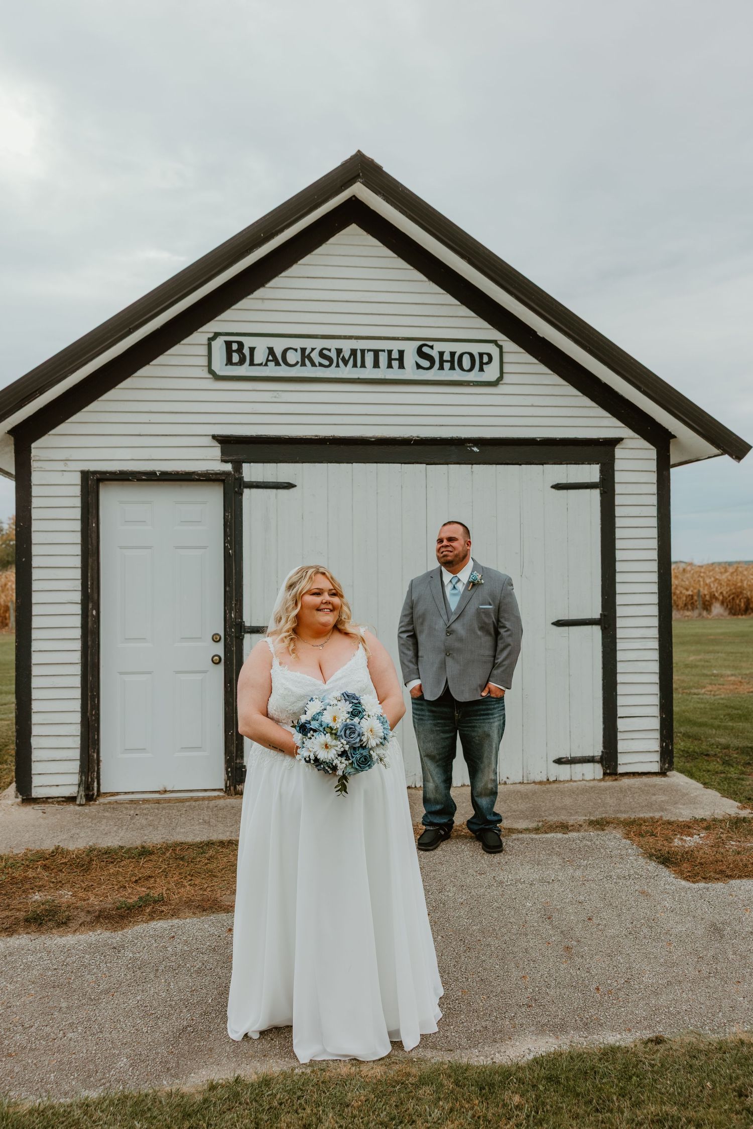 Bride and groom pose in front of a white Blacksmith Shop building at their rustic wedding venue. Northwest Indiana wedding venues.