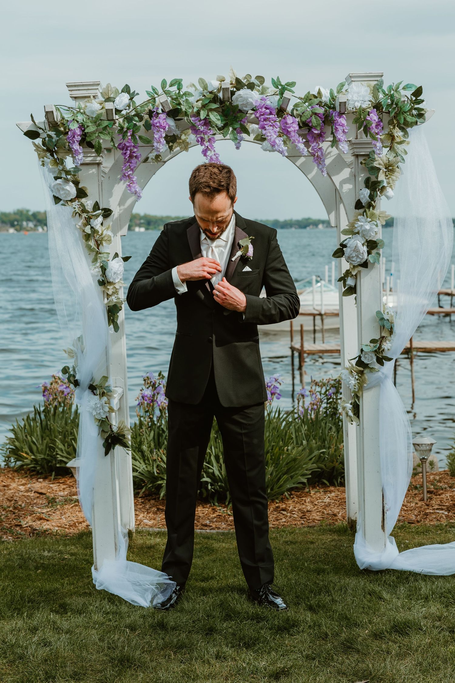 Dapper groom in black suit adjusts boutonniere under a purple floral arch by the waterfront.