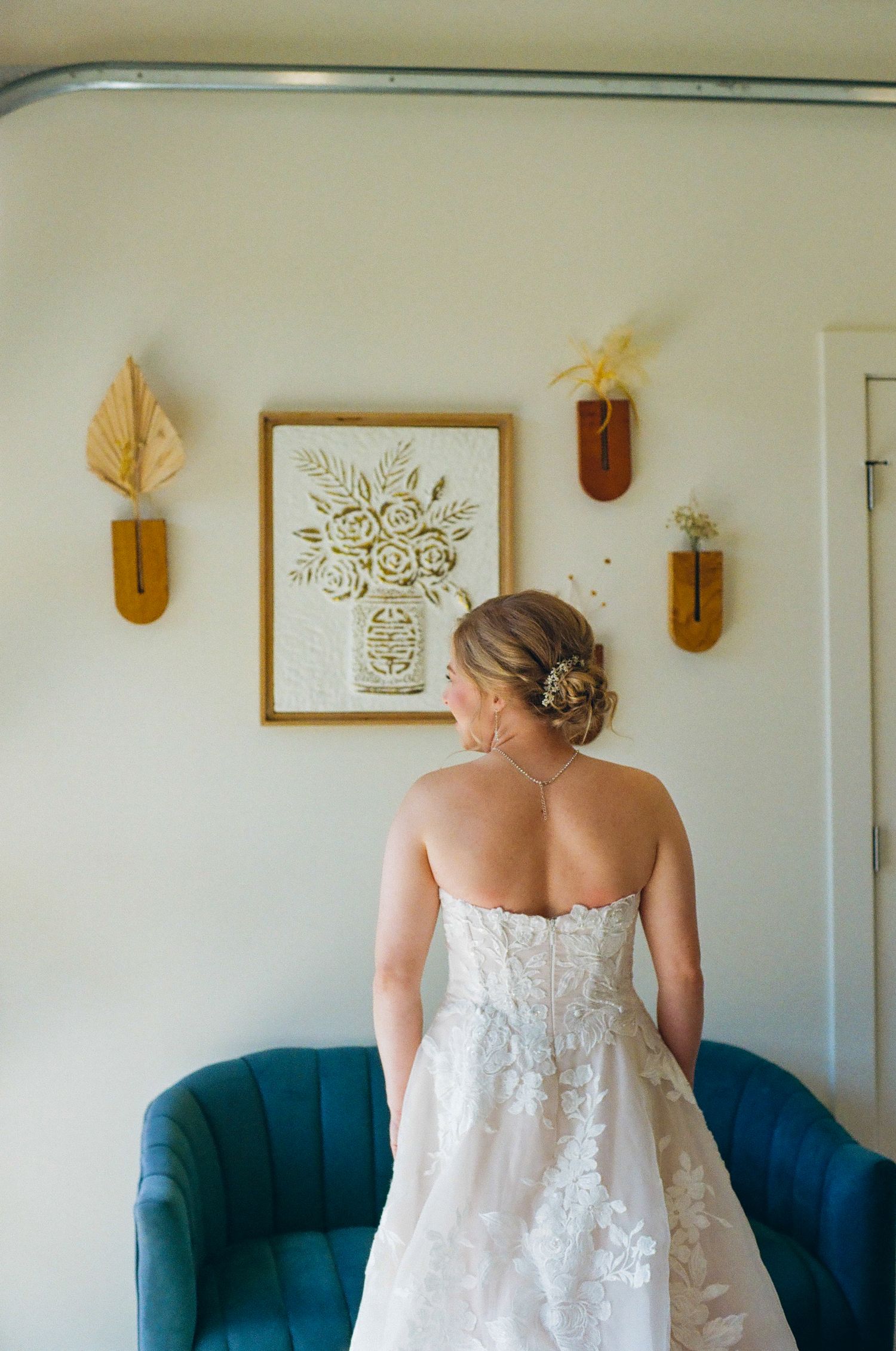 Bride in strapless lace wedding gown viewed from behind, standing near teal velvet chair with floral wall art.