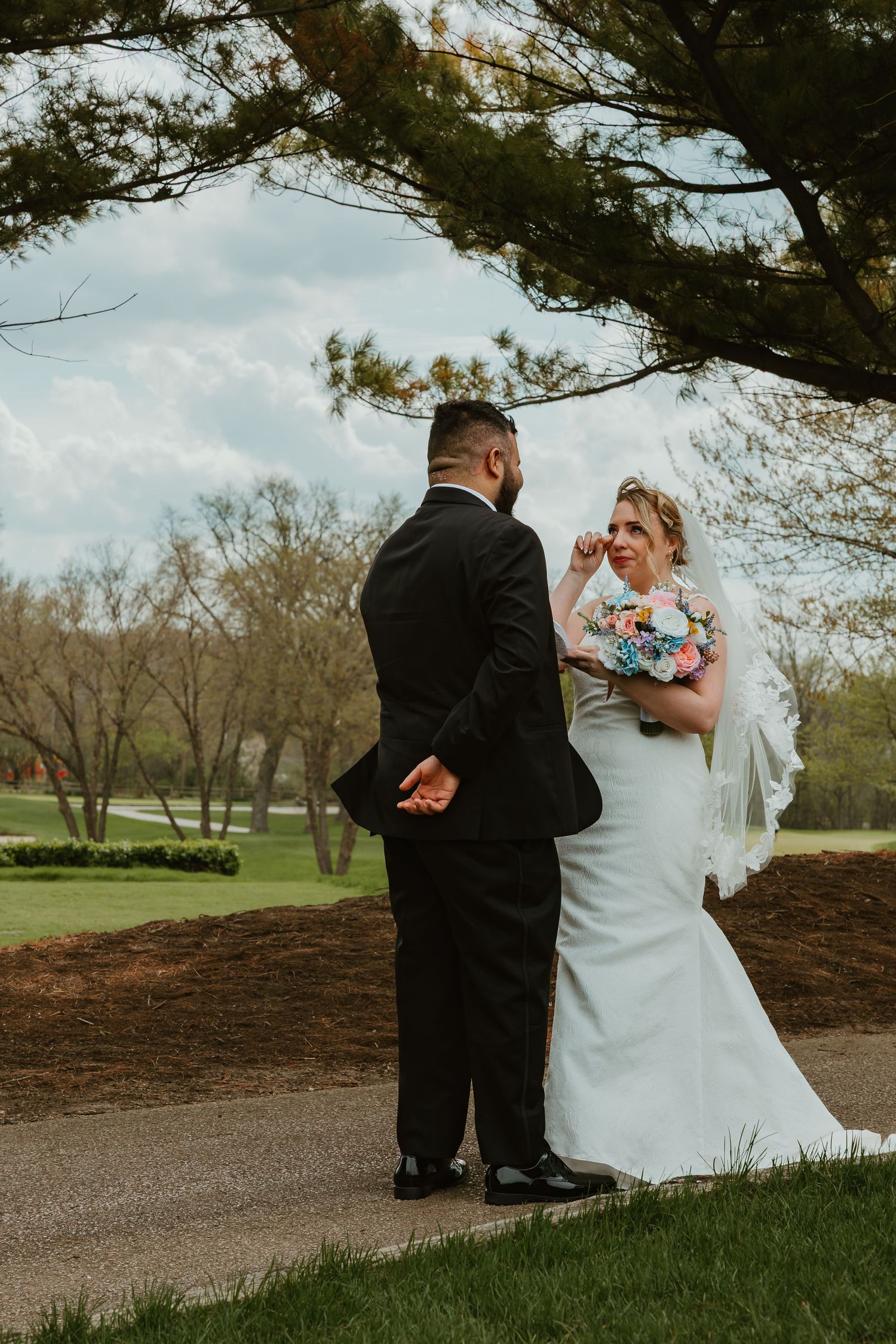 Emotional bride wiping tears during first look moment with groom in black tuxedo on golf course path. Chicagoland wedding venues.
