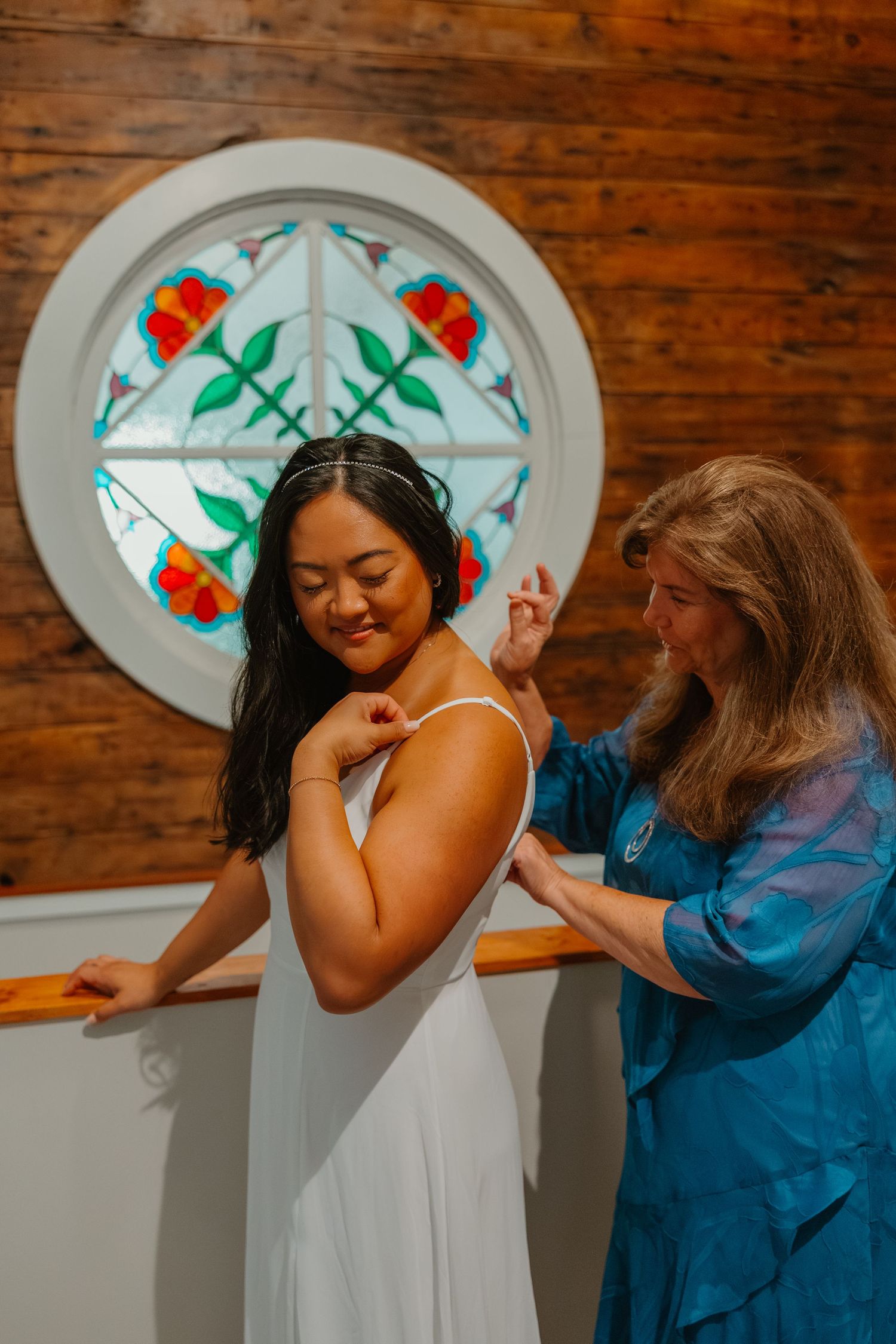 Bride in white gown gets dress adjusted by older woman near a colorful stained glass window. Michigan elopement.