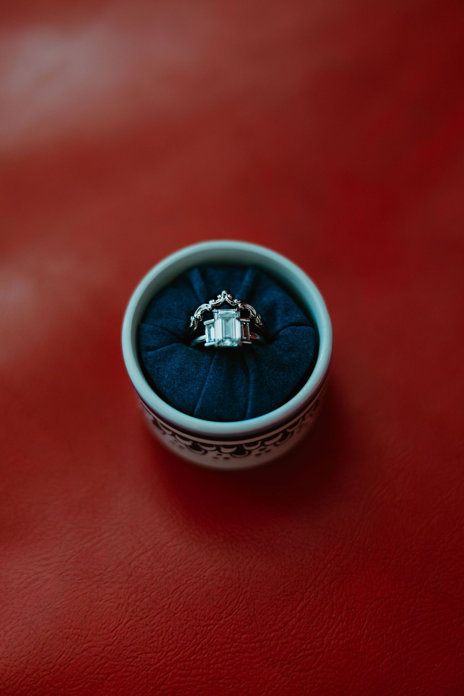 Diamond engagement ring with ornate setting displayed in a round blue velvet ring box on red leather.