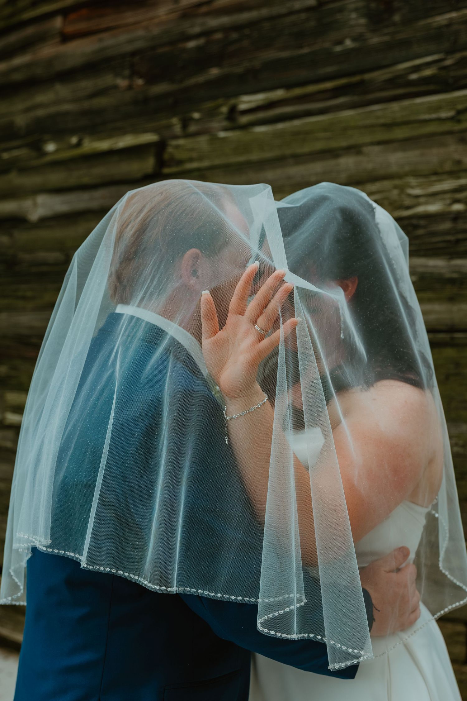 Bride and groom share a romantic kiss hidden beneath the bride's flowing white veil against a rustic wood backdrop. Northwest Indiana weddin