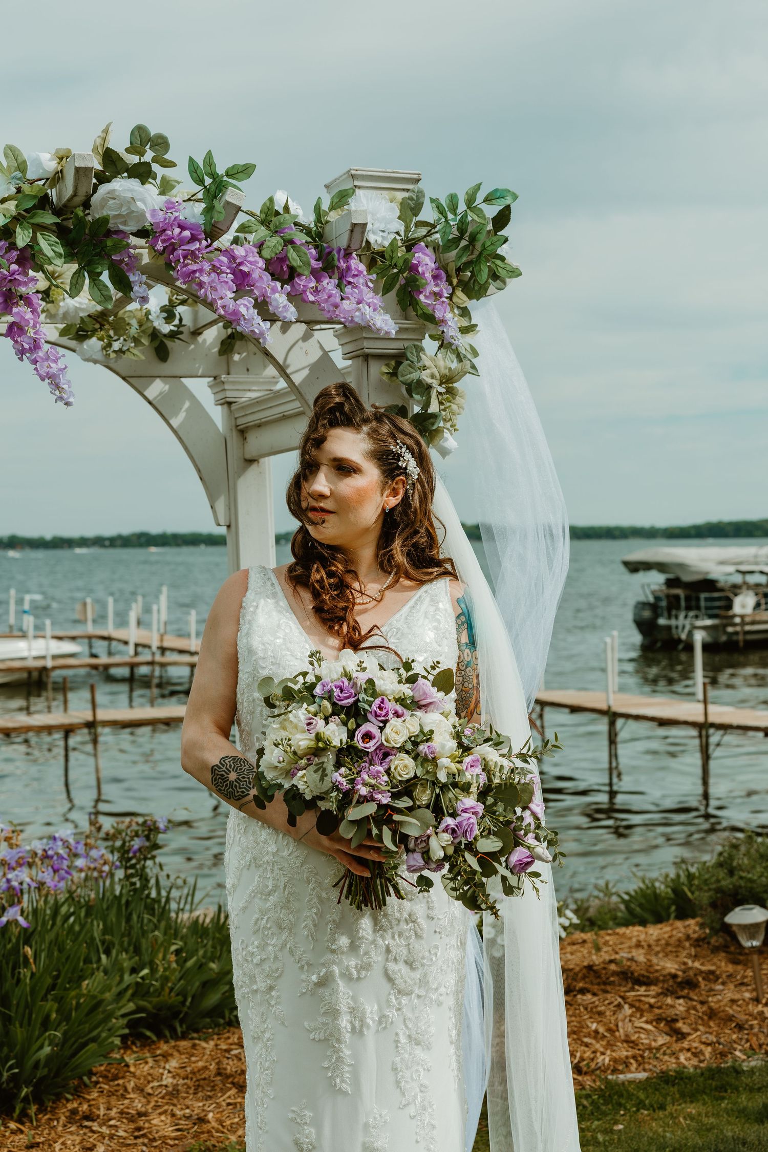 Bride in lace gown holds purple floral bouquet under floral arch by waterfront.