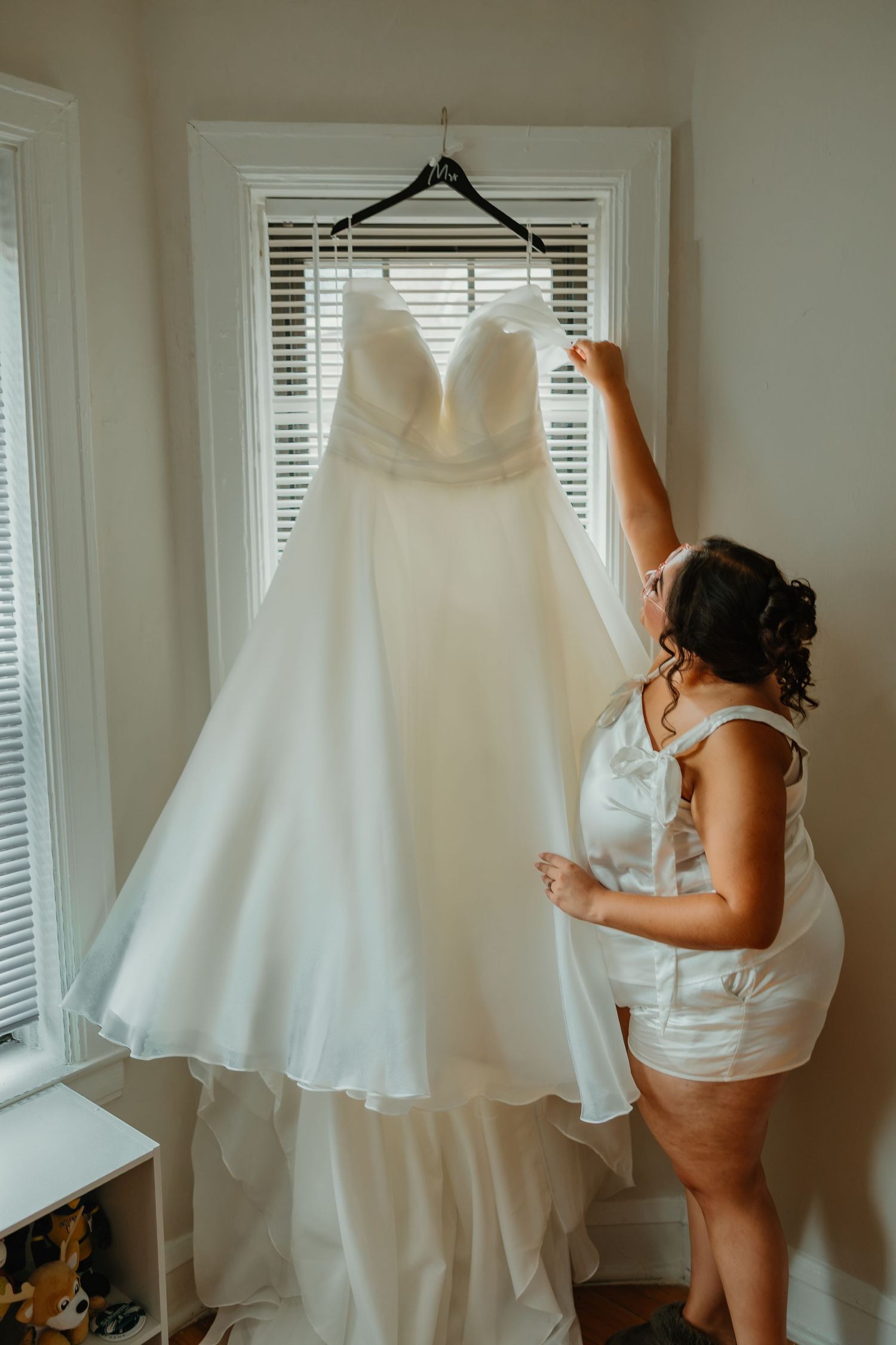 Bride in white satin romper reaches up to touch her elegant strapless wedding gown hanging in window. Chicagoland weddings.