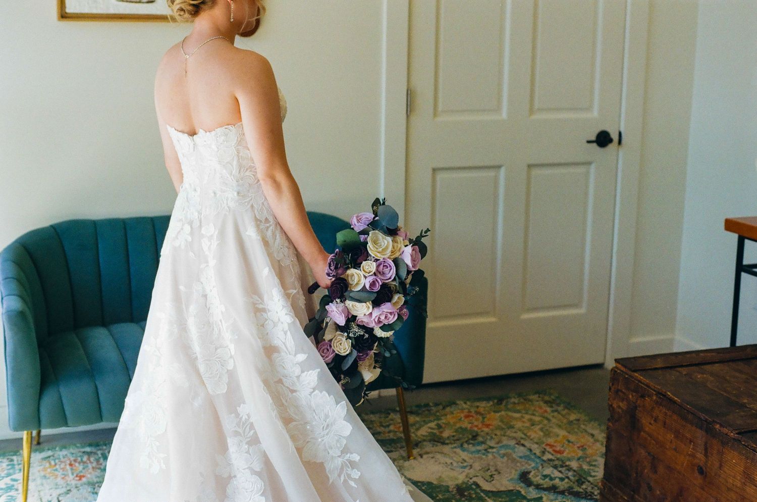 Bride in strapless white lace wedding gown holding purple floral bouquet, standing near teal chair and white door.