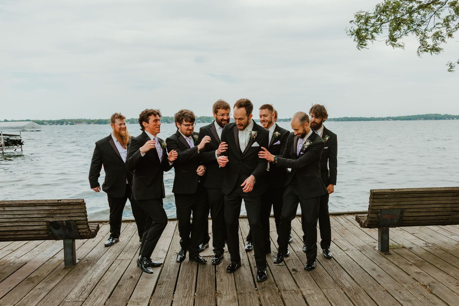 Groom and groomsmen laughing together on a wooden dock by the lake in black suits.