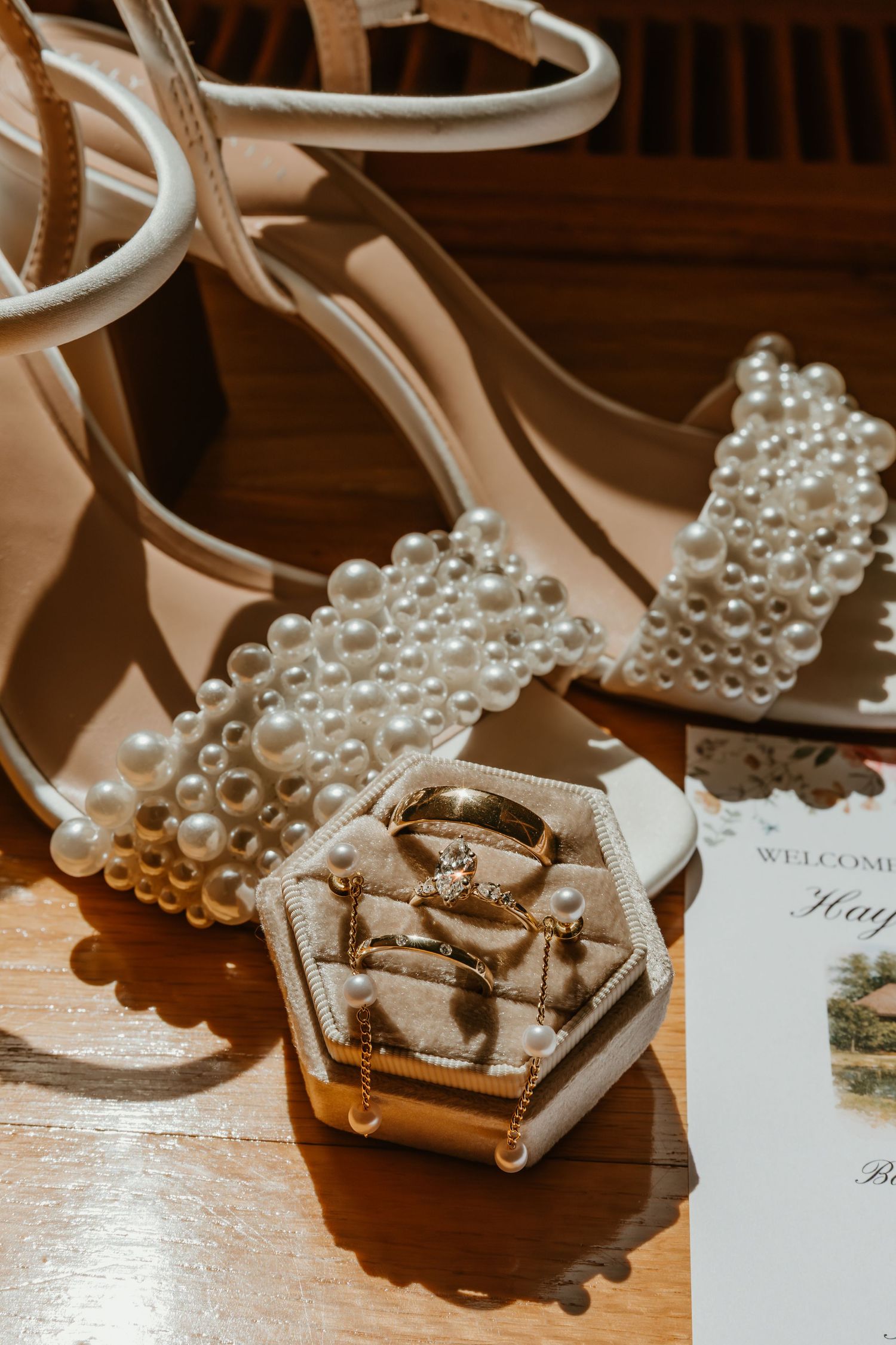 Pearl-embellished bridal heels beside hexagonal ring box with gold wedding bands on wood.