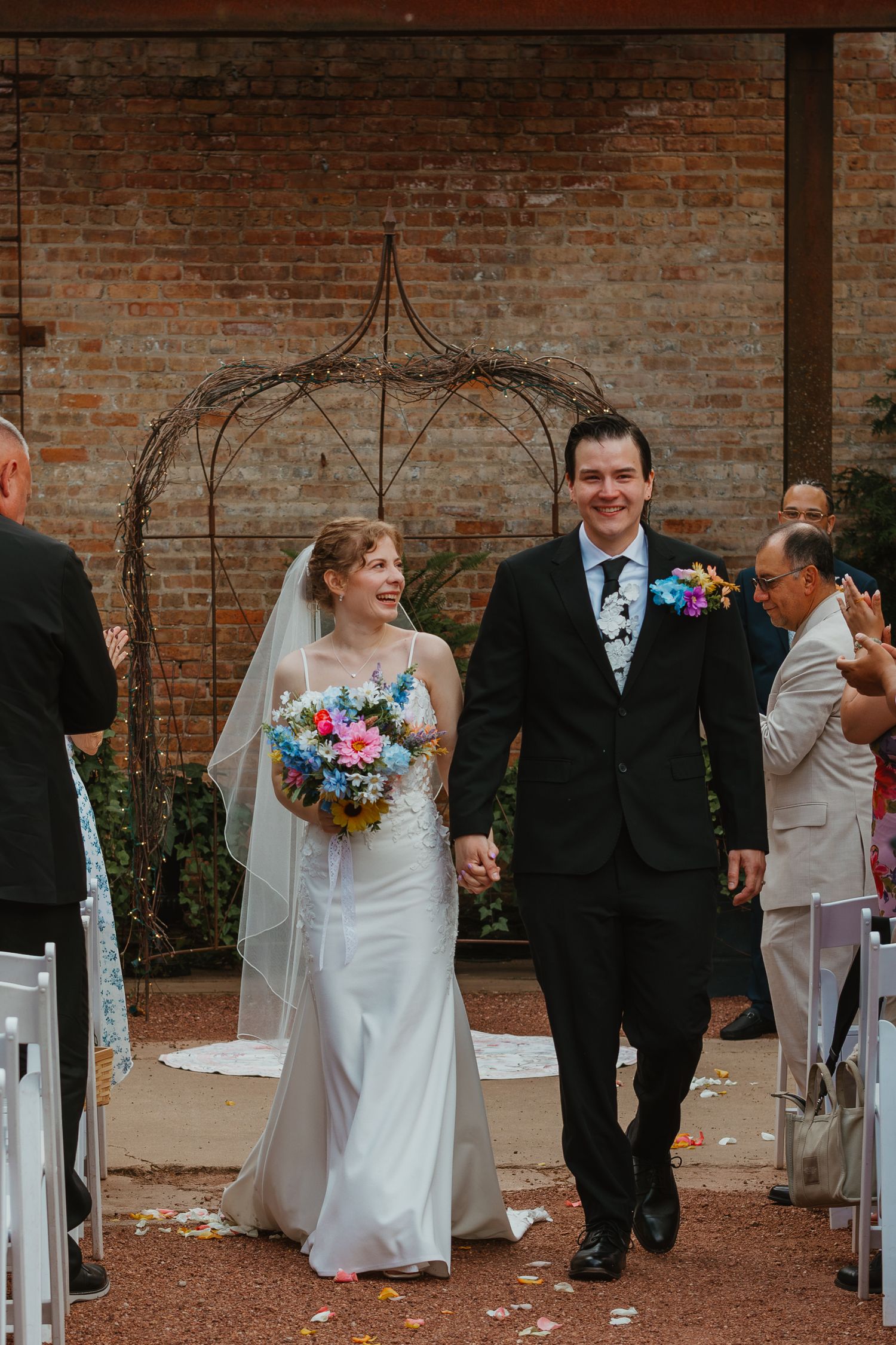 Newlyweds walk down aisle smiling, bride holding vibrant bouquet, groom in black suit, brick wall backdrop. Chicagoland wedding venues.
