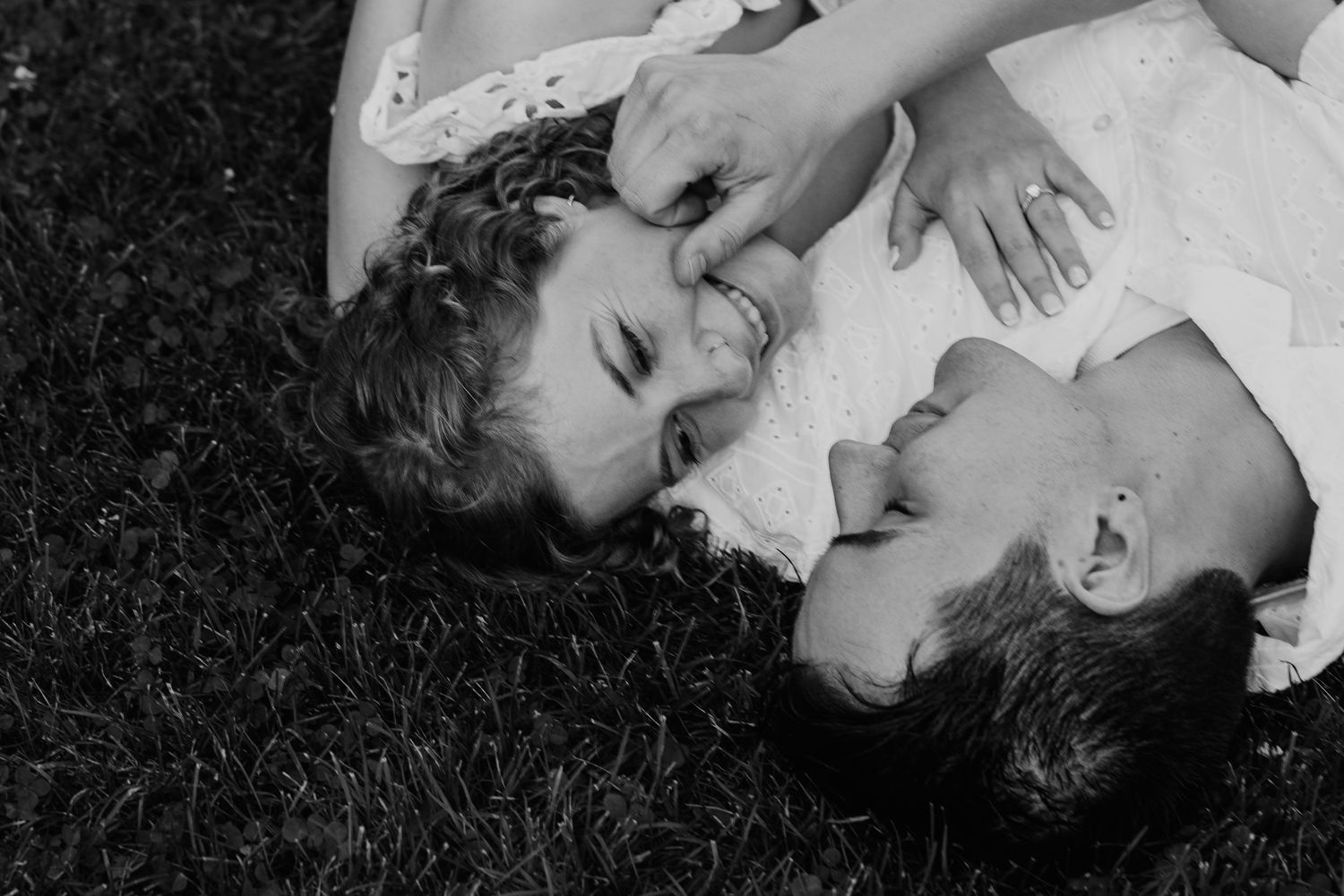 Black and white photo of bride and groom lying on grass face to face laughing together. Chicagoland engagement photos.