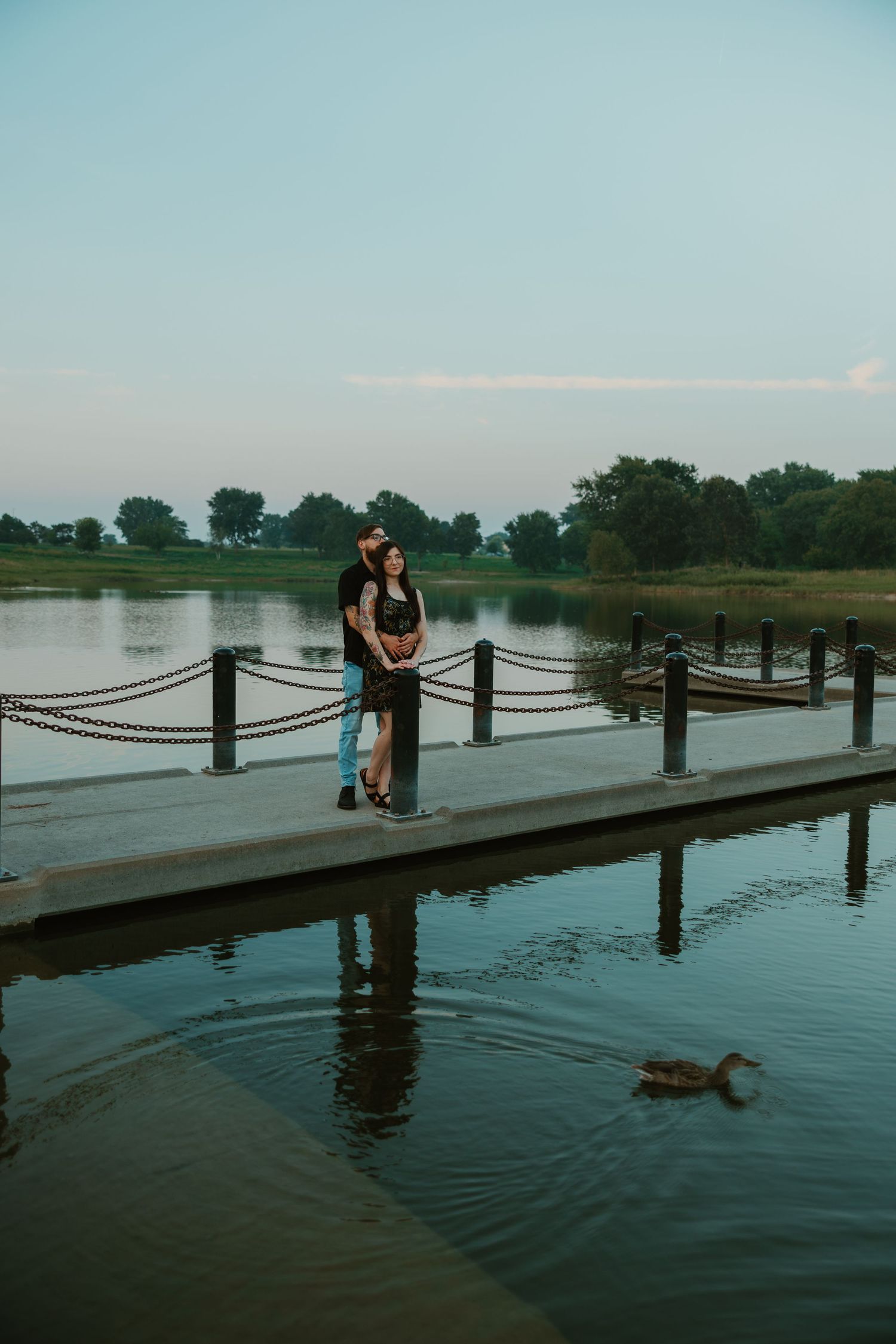 Couple embracing on a wooden dock by a calm lake at dusk, with a duck swimming nearby. Chicagoland engagement photos.