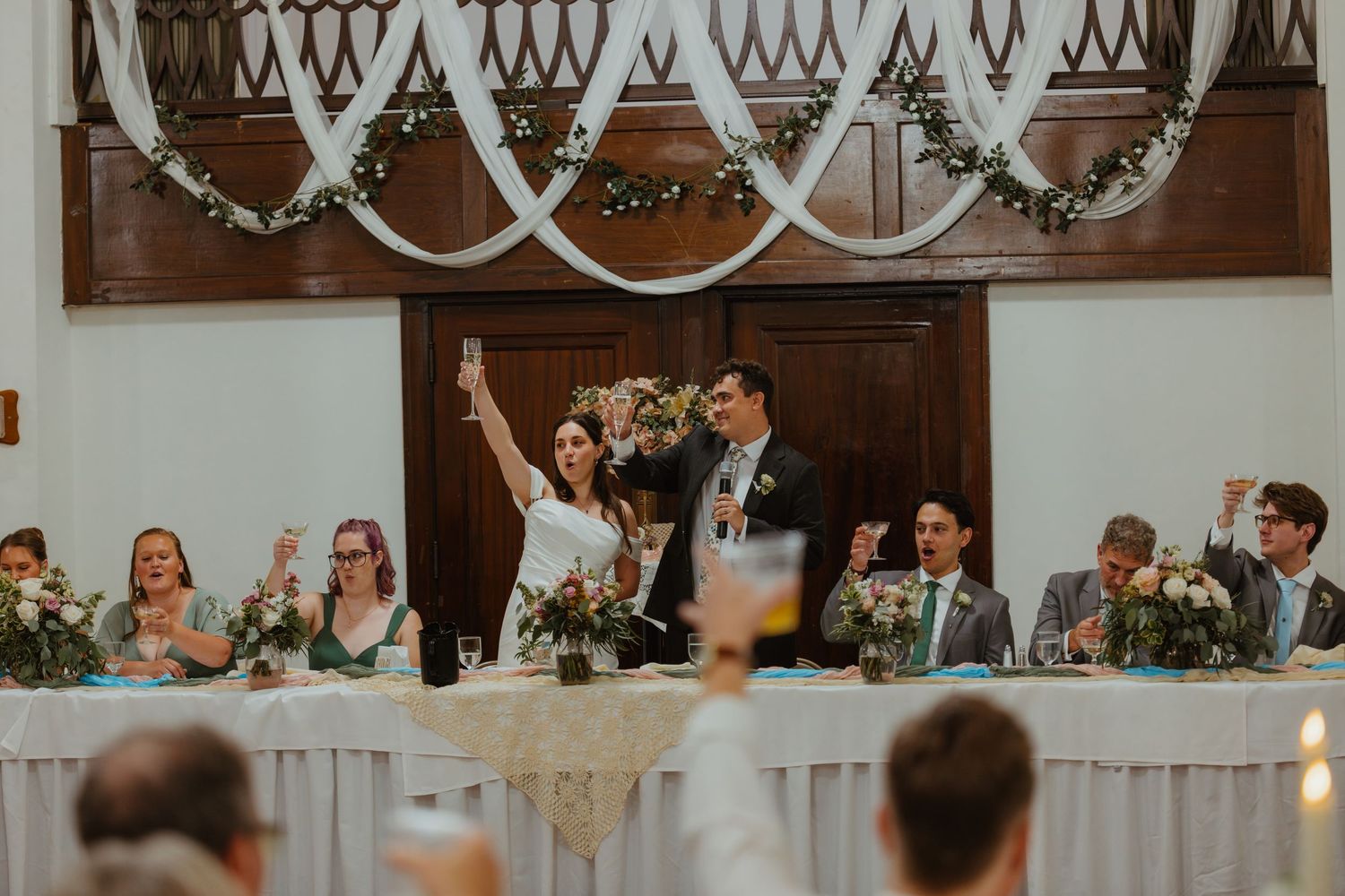 Wedding party raises glasses in a toast at the head table in an elegantly decorated reception hall.