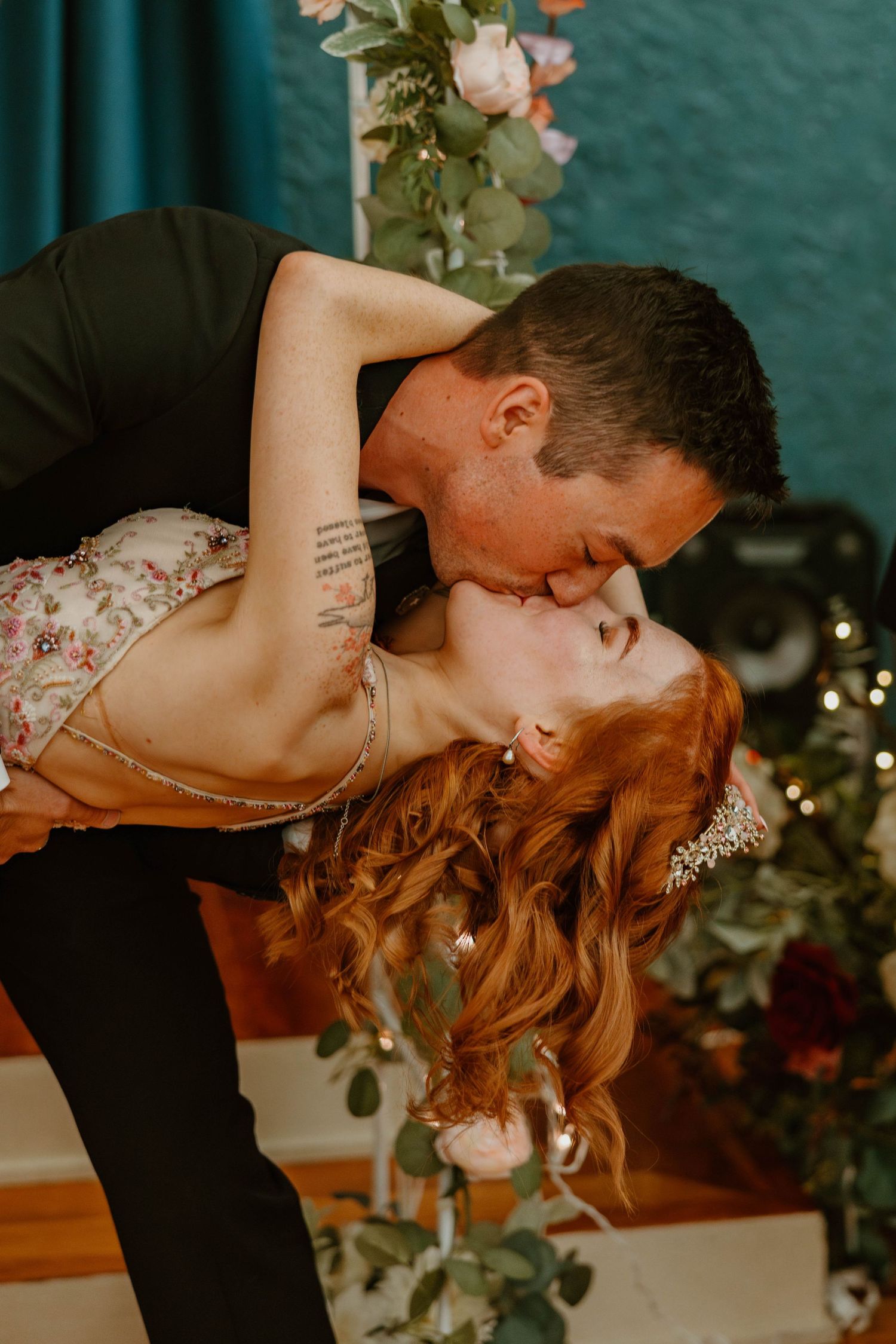 Groom dips and kisses red-haired bride in floral dress surrounded by wedding flowers.
