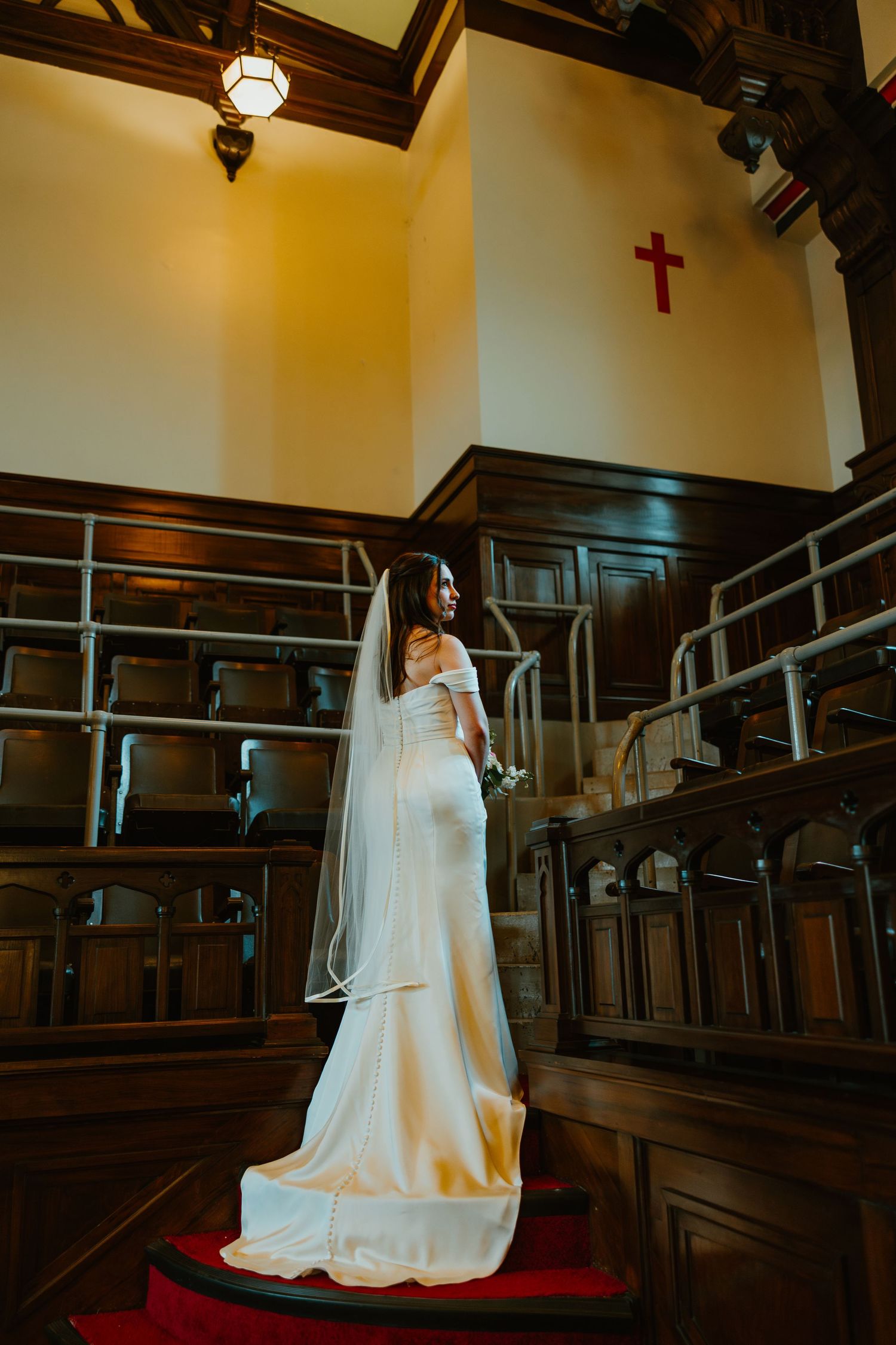 Elegant bride in white off-shoulder gown with cathedral veil stands on red-carpeted church stairs.