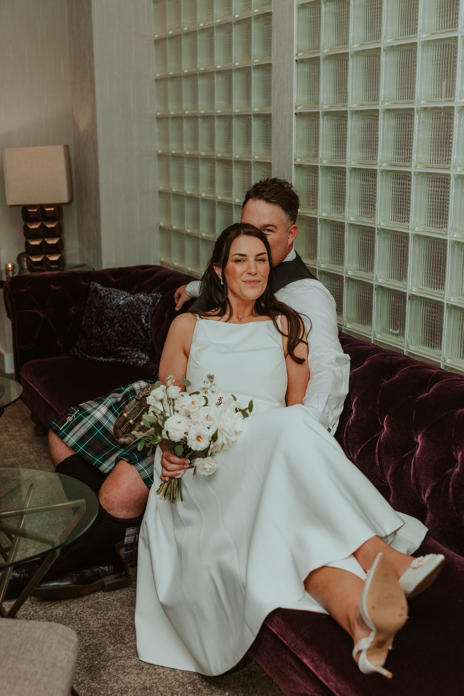 Bride and kilt-wearing groom relax on velvet sofa with bouquet. Chicago Thompson Hotel wedding.