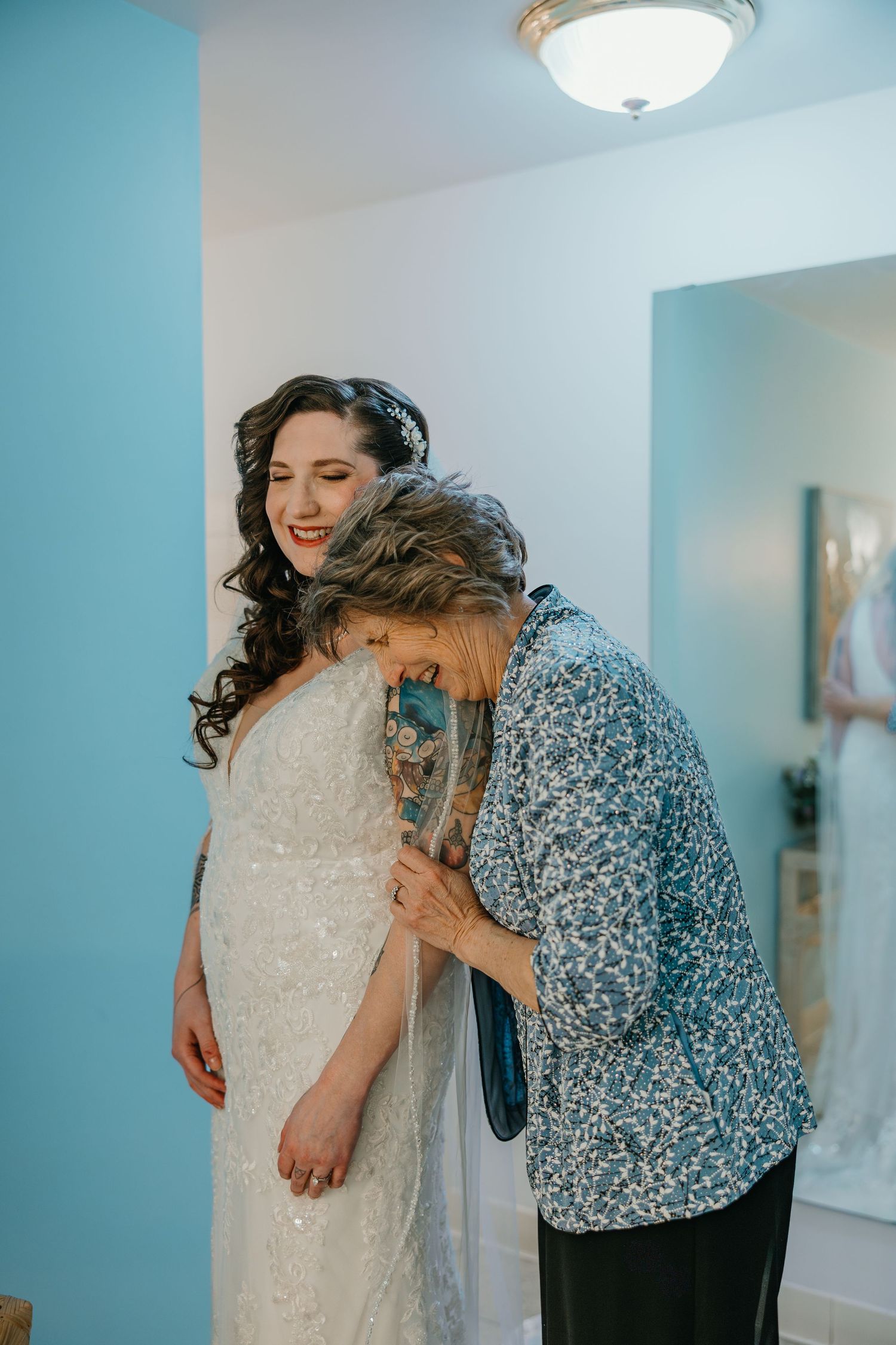 Elderly woman lovingly embraces smiling bride in beaded white gown during wedding preparation.