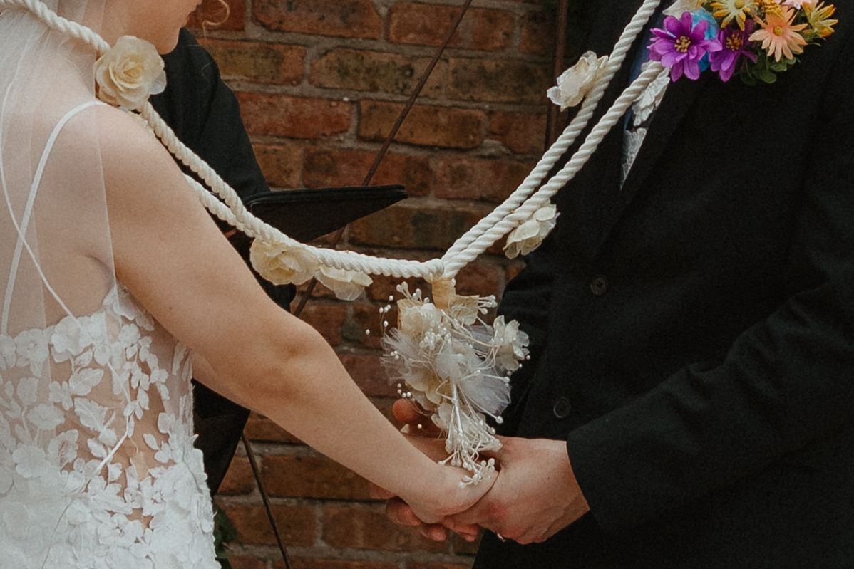 Bride and groom participate in rope handfasting ceremony, holding hands bound with white rope at brick wall. Chicagoland wedding venues.