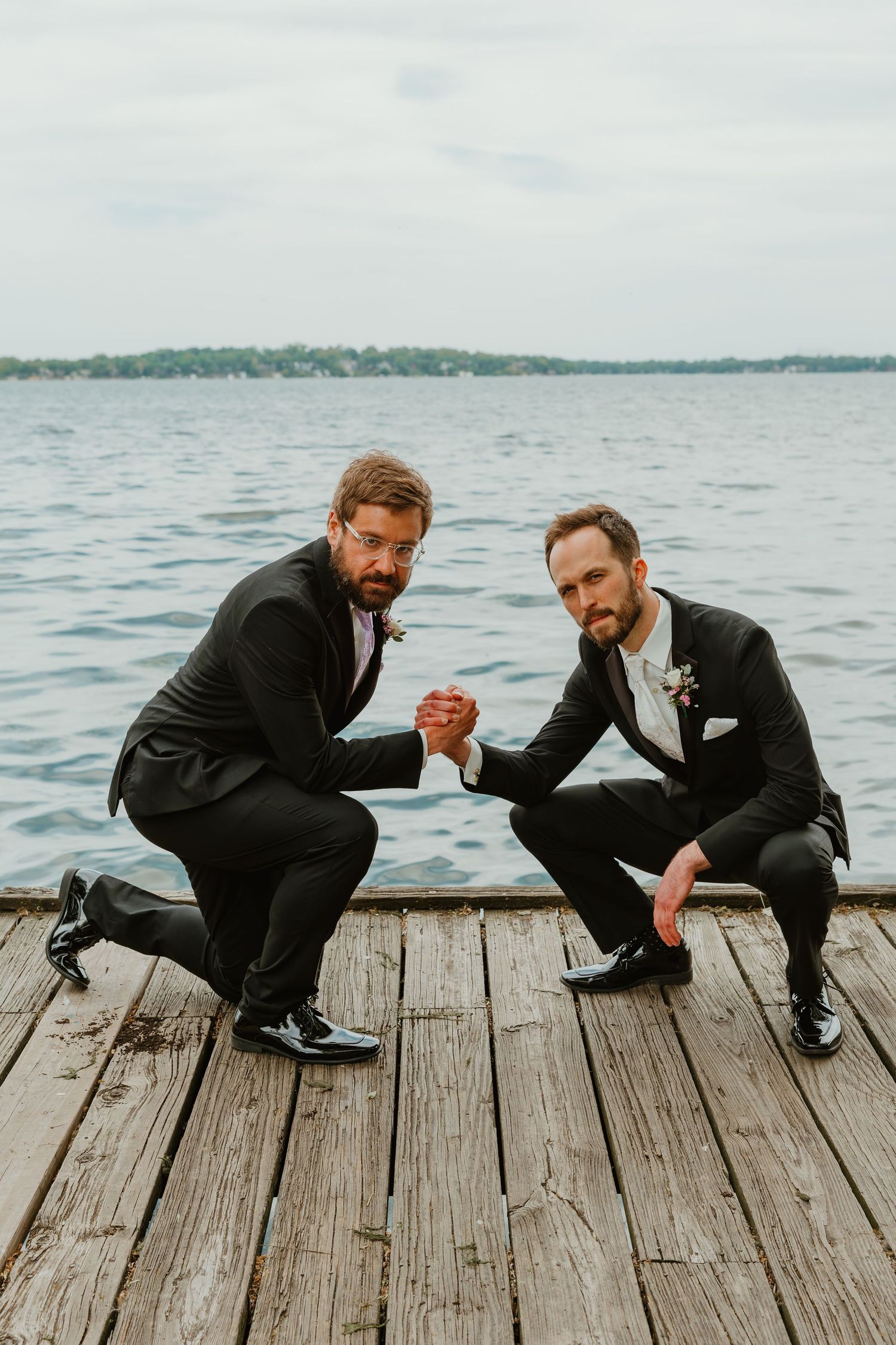 Two men in black suits posing with clasped hands on wooden dock with lake in background.