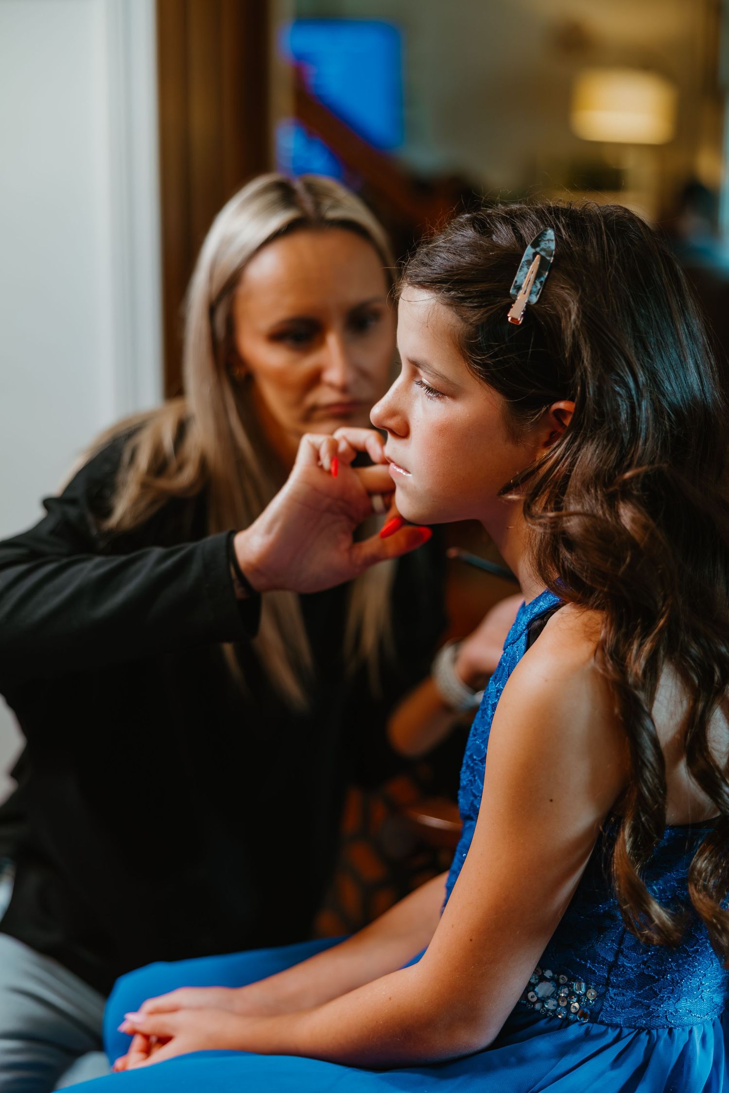 Blonde woman applies lip color to young girl in blue dress during wedding day preparations. Northwest Indiana wedding venues.