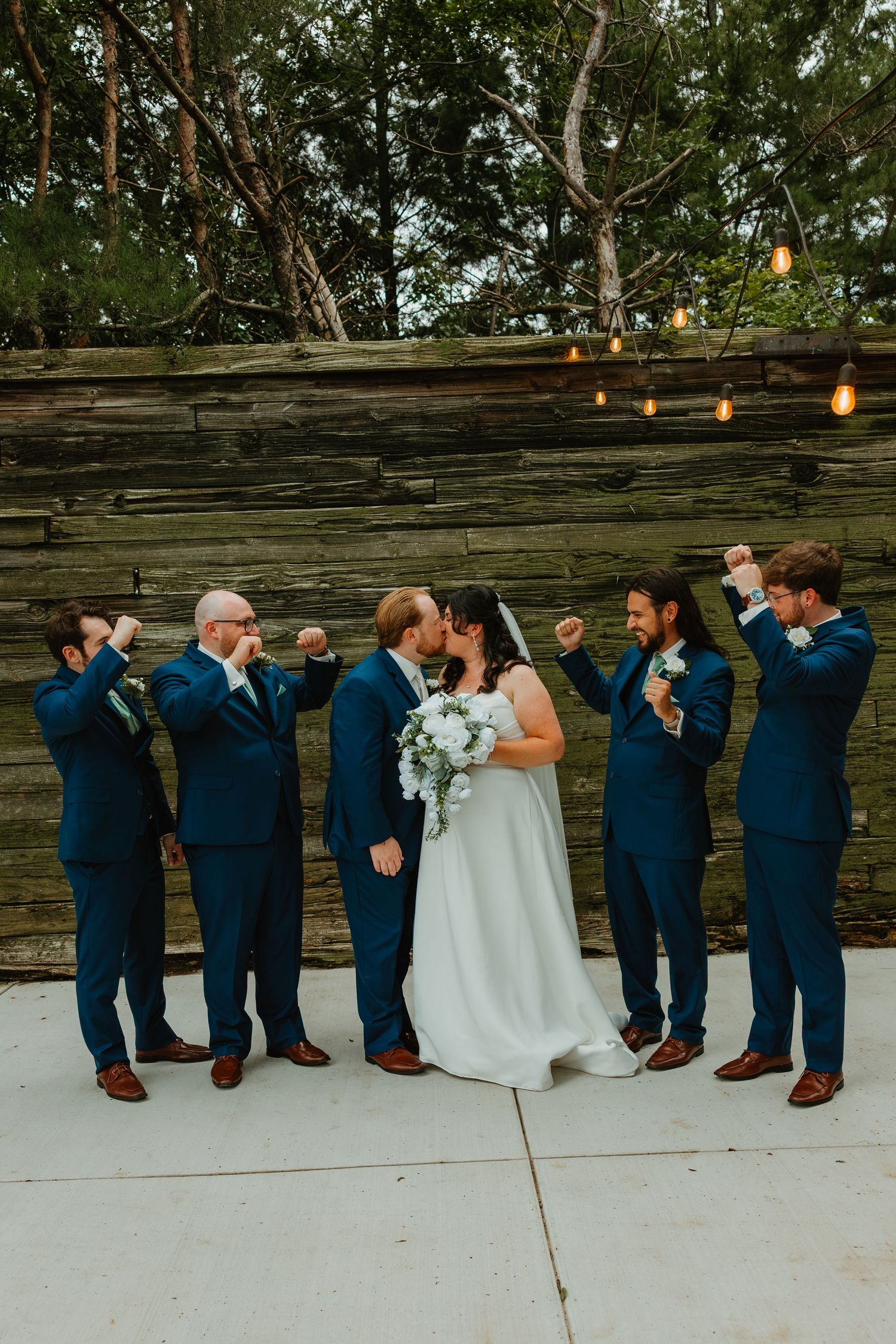 Groom kisses bride while groomsmen in navy suits celebrate with raised fists at outdoor wedding. Northwest Indiana wedding venues.