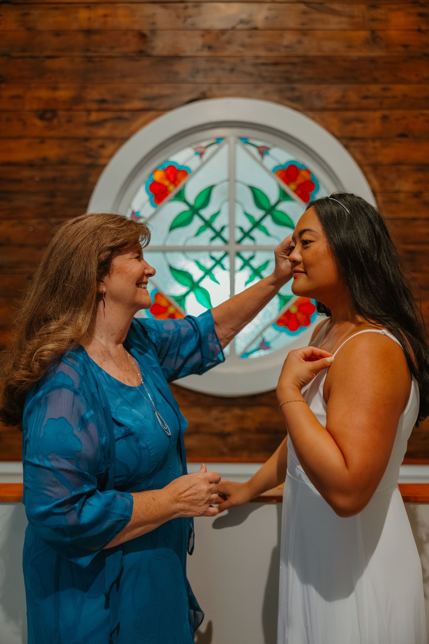 Two women sharing a tender moment in front of a colorful stained glass window in a rustic wooden chapel. Michigan elopement photos.