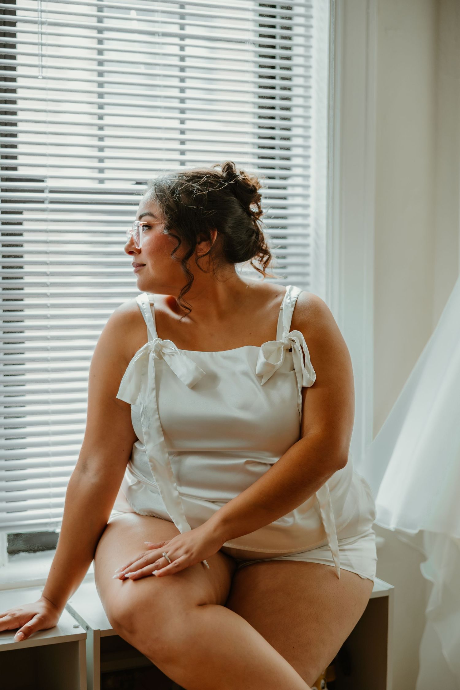 Bride in white satin bridal romper sits thoughtfully by window with soft natural light on wedding morning. Chicagoland weddings.