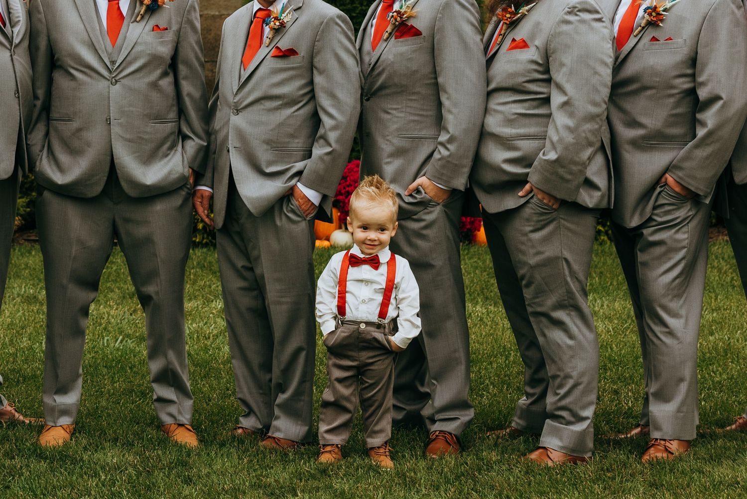Adorable toddler ring bearer in suspenders and bow tie stands between groomsmen in gray suits.
