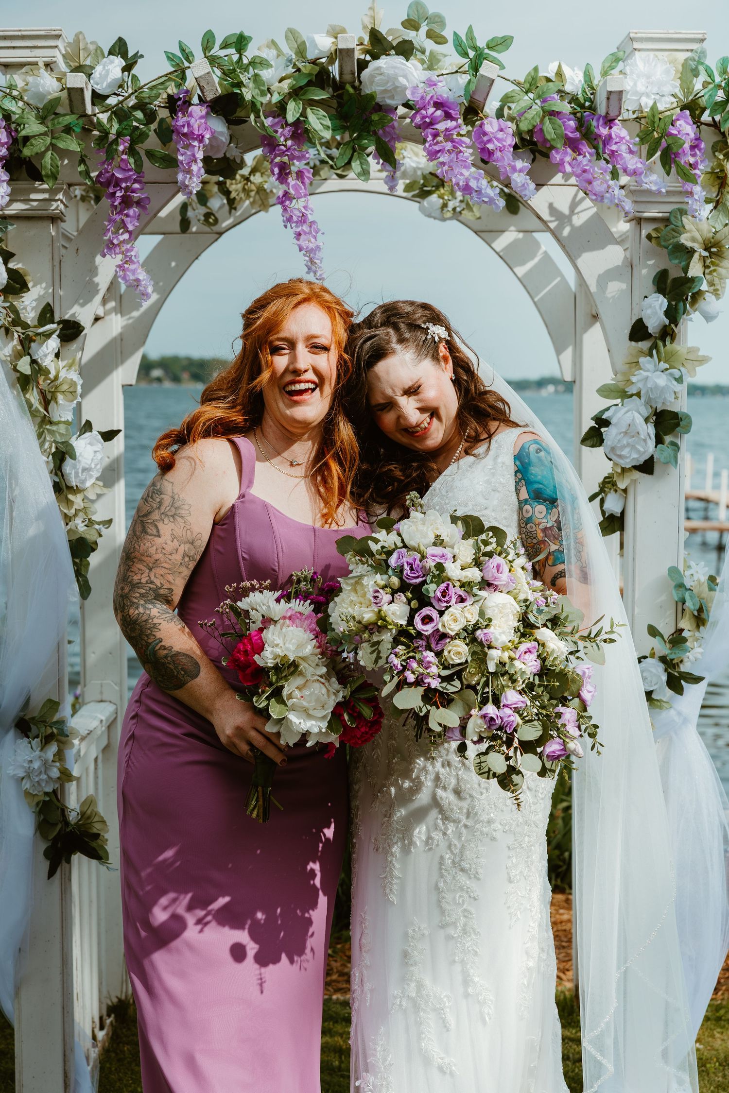 Bridesmaid in purple gown and bride in lace dress smile under floral arch.