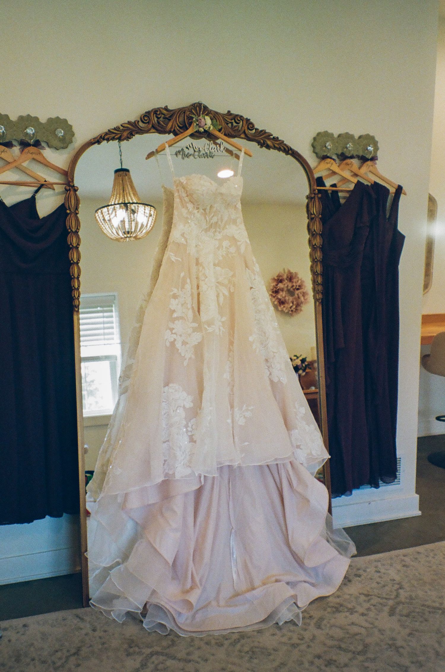 Elegant blush wedding gown with lace appliques hanging on ornate gold mirror, flanked by dark bridesmaid dresses.