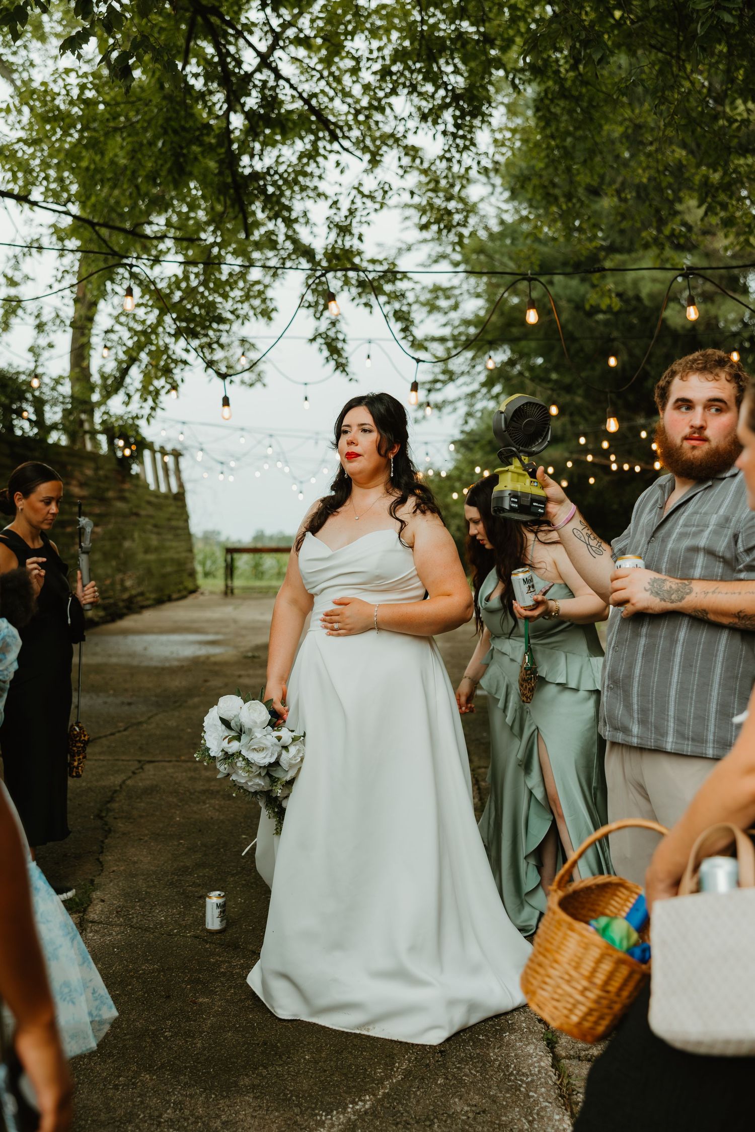 Bride in white strapless gown walks down outdoor aisle holding white bouquet with string lights above. Northwest Indiana wedding venues.