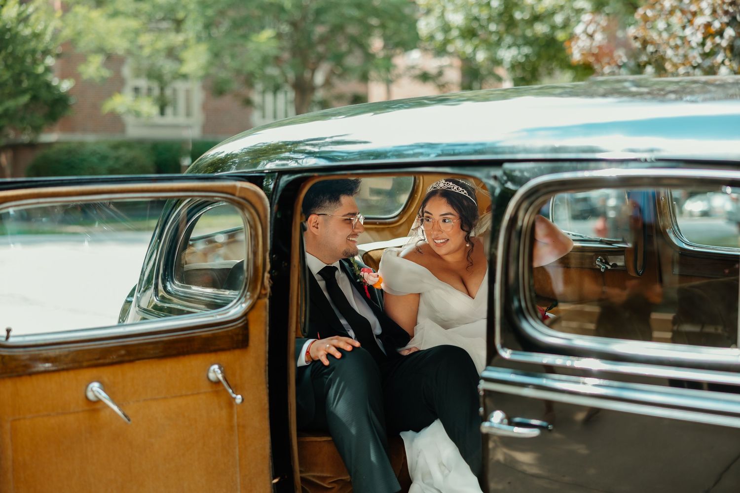 Bride and groom share a playful moment seated in doorway of a classic vintage wedding car. Chicagoland weddings.