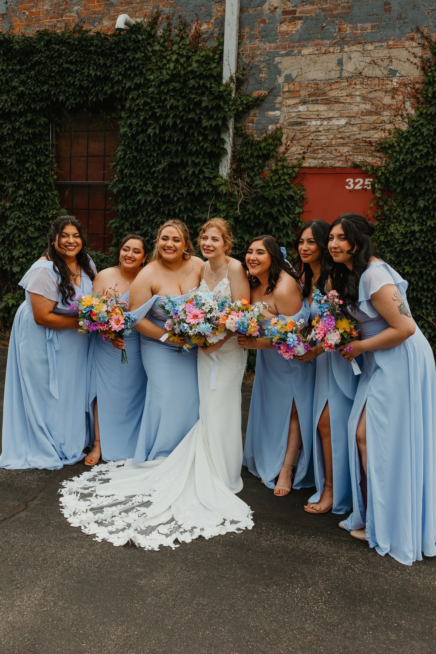 Bride in lace gown with bridesmaids in blue dresses holding colorful bouquets outdoors. Chicagoland wedding venues.