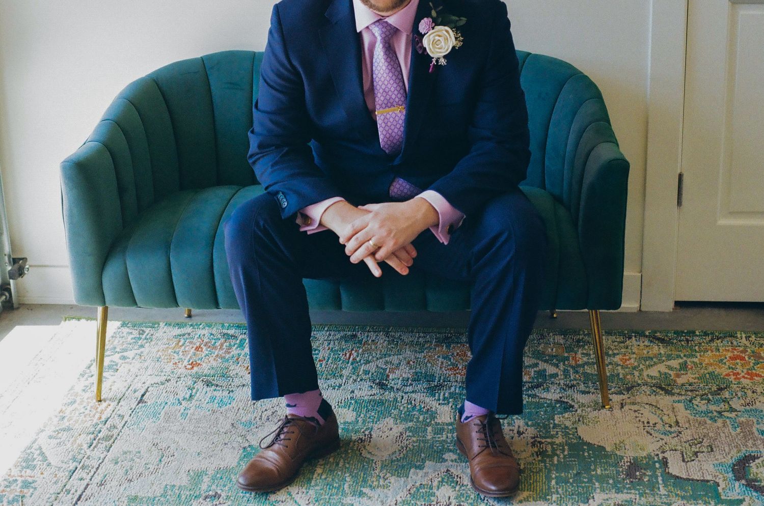 Groom in navy suit with pink tie and purple socks sits on teal velvet sofa, wearing brown oxford shoes.
