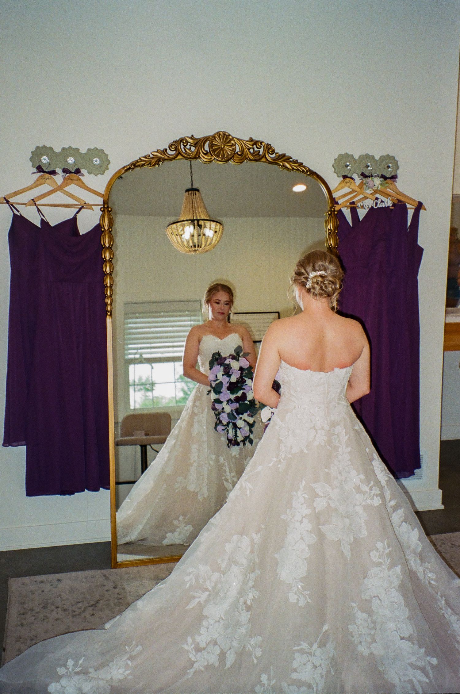 Bride in lace ballgown views reflection in ornate gold mirror, purple bridesmaid dresses hanging on either side.