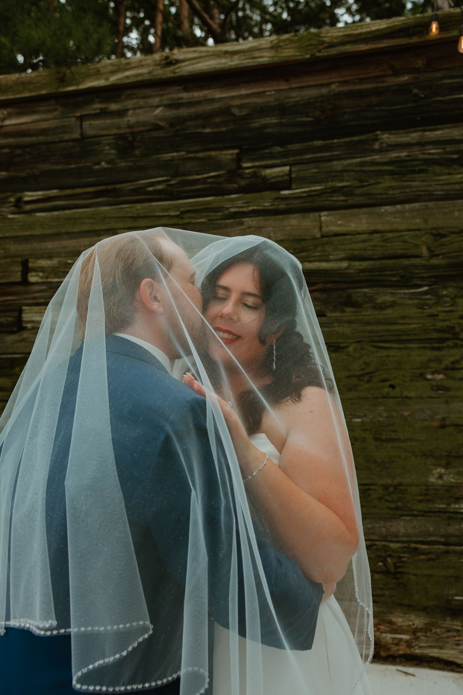 Bride and groom share intimate moment under sheer wedding veil against rustic wooden wall backdrop. Northwest Indiana wedding venues.