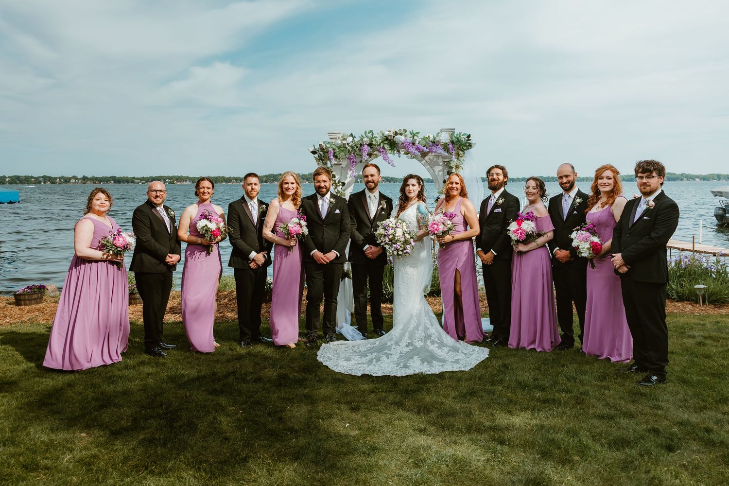 Wedding party poses in purple and black attire by lakeside floral arch.