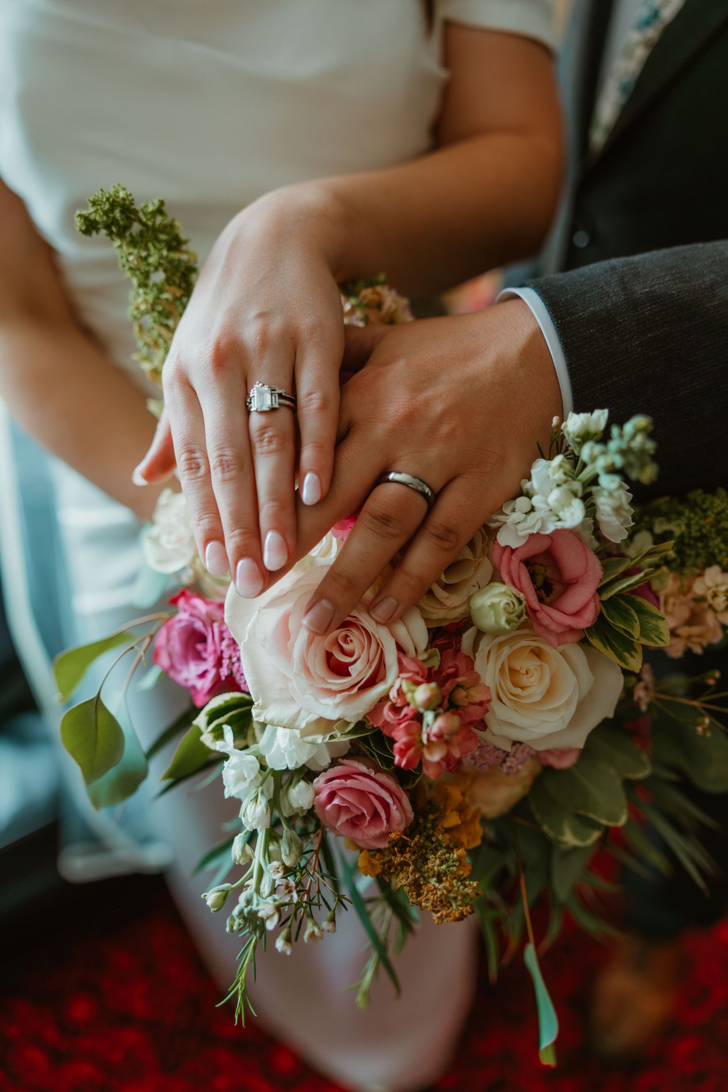 Bride and groom display wedding rings while hands rest on a colorful bridal bouquet of pink roses.