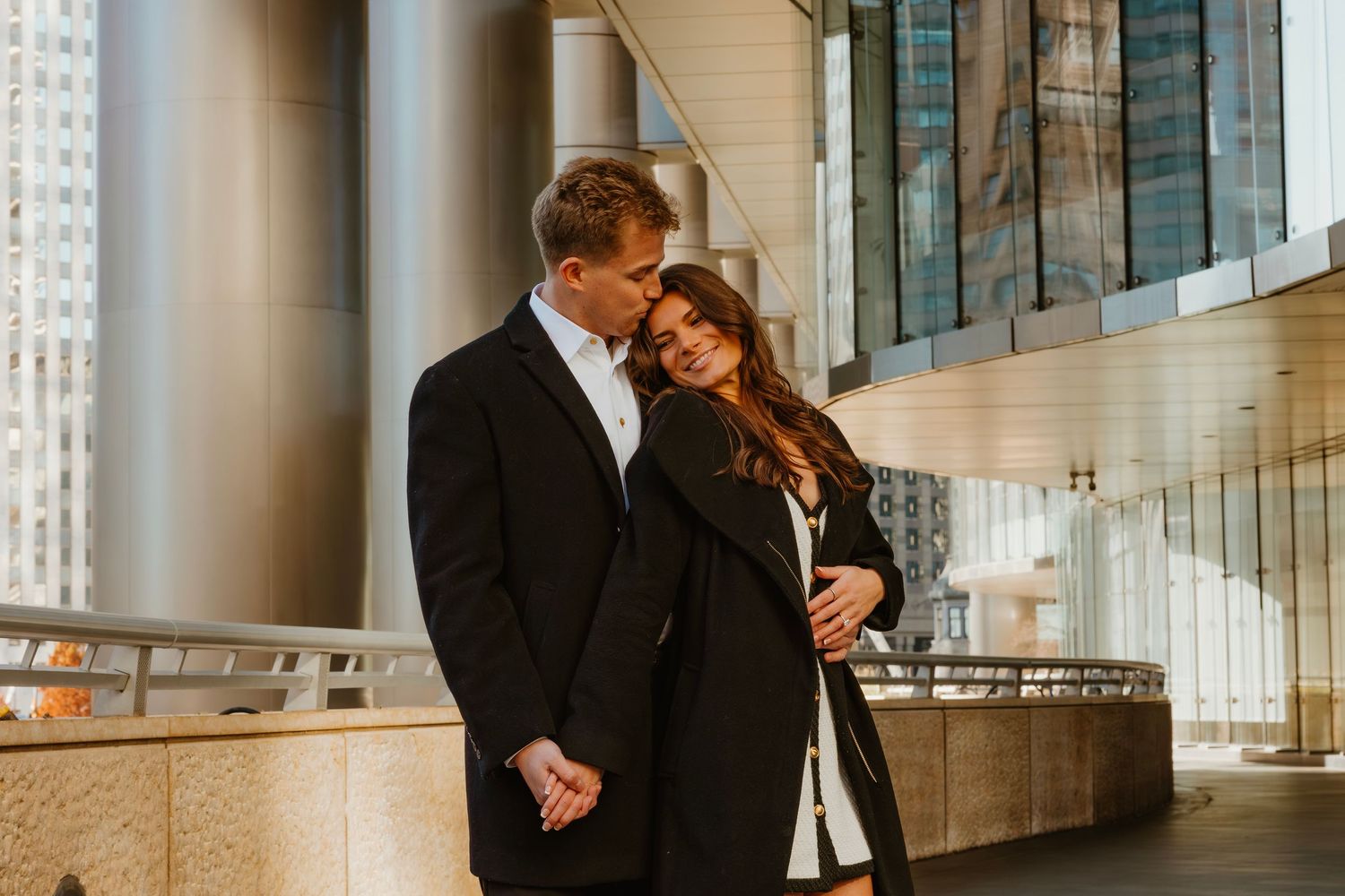 Couple cuddling close together outside a modern glass building in a city setting. Outdoor Chicago engagement photos.