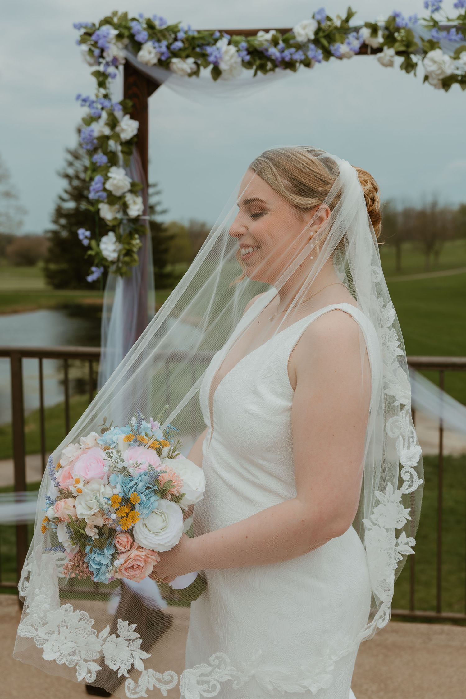 Smiling bride in white gown and lace veil holding pastel bouquet beneath a floral wedding arch outdoors. Chicagoland wedding venues.