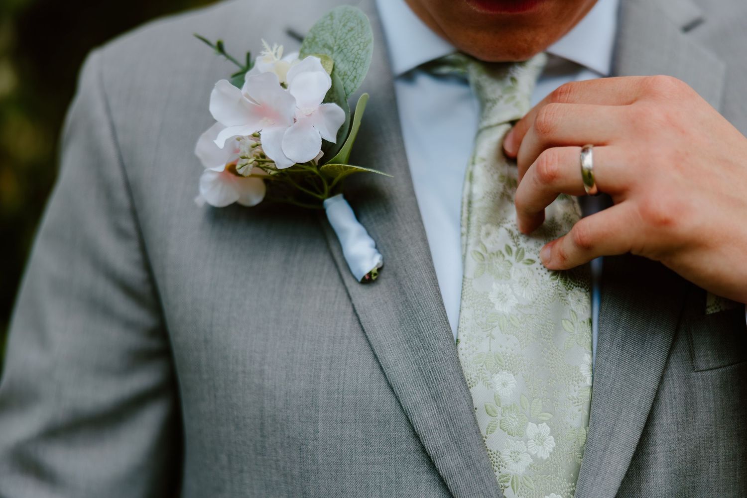 Groom in grey suit adjusting floral tie with pink boutonniere and wedding band detail. Northwest Indiana event center wedding photos.